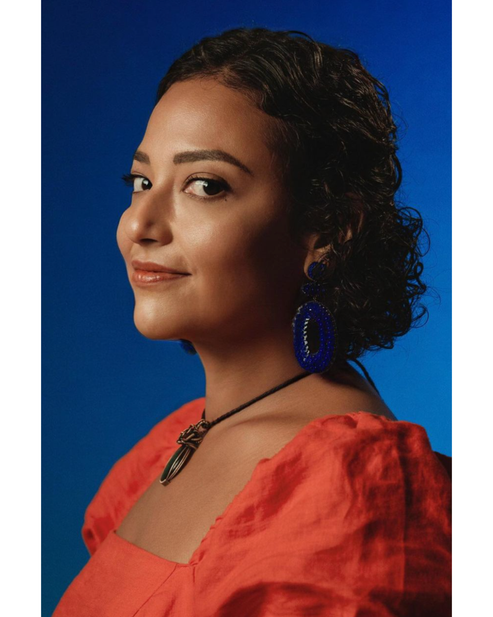 Portrait of a woman with short curly dark hair against a blue background, wearing large blue earrings, a black necklace with a pendant, and a red blouse with puffed sleeves.
