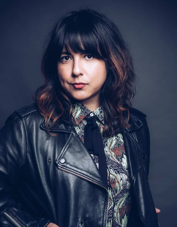A woman with shoulder-length dark hair with brown highlights, wearing a black leather jacket over a patterned shirt, against a dark background.