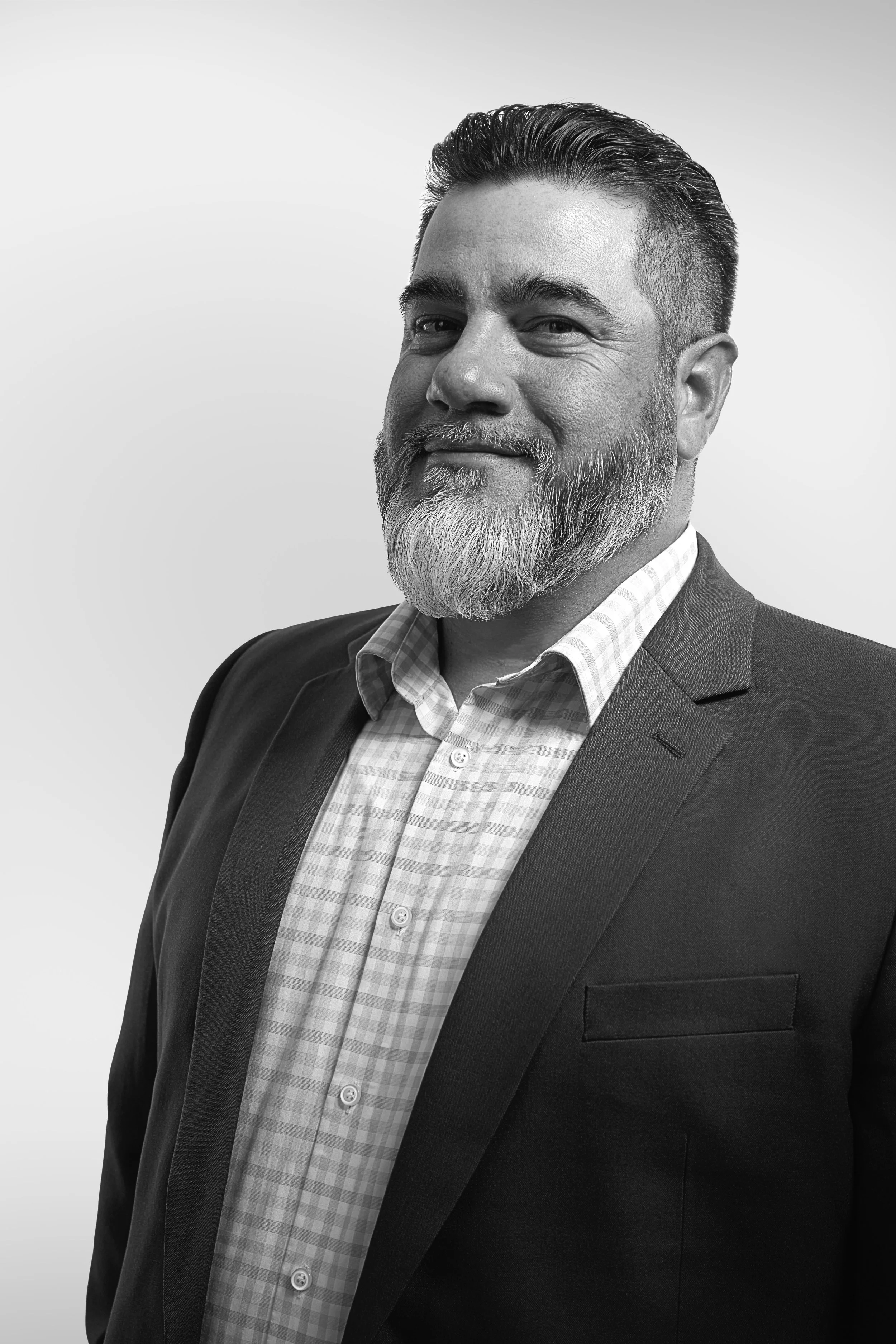 Black and white portrait of a middle-aged man with a beard and mustache, wearing a suit jacket and a checkered shirt, smiling slightly, against a light gradient background.