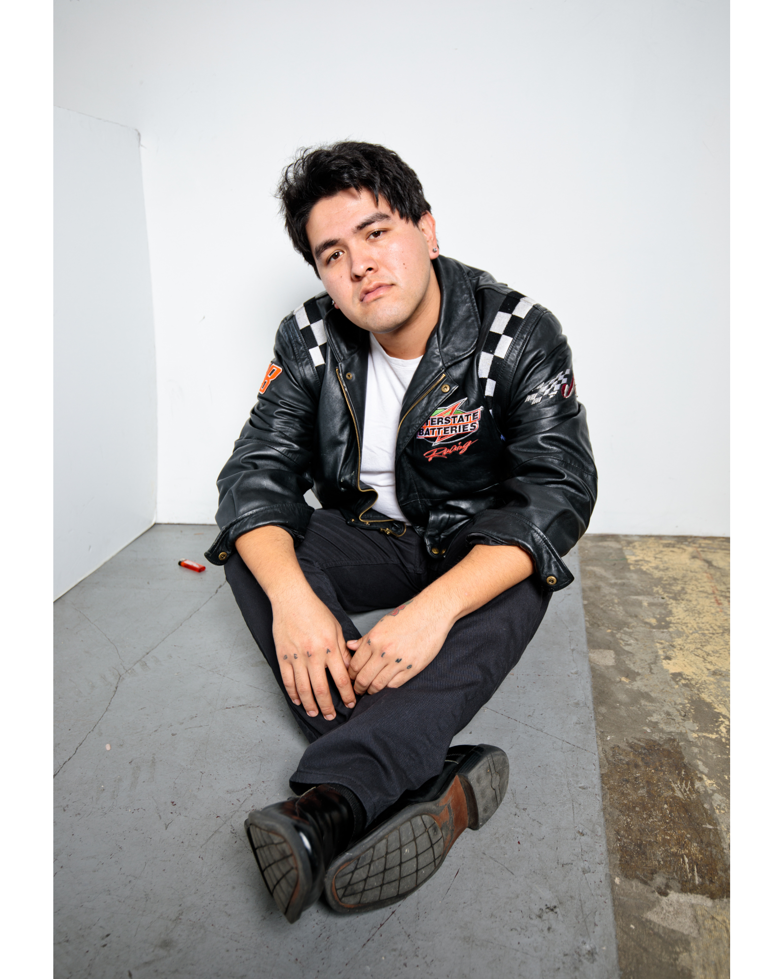 A young man with dark hair sitting on the floor against a white wall, wearing a black leather racing jacket with patches and checkered details, black pants, and black boots, with a relaxed and slightly serious expression.