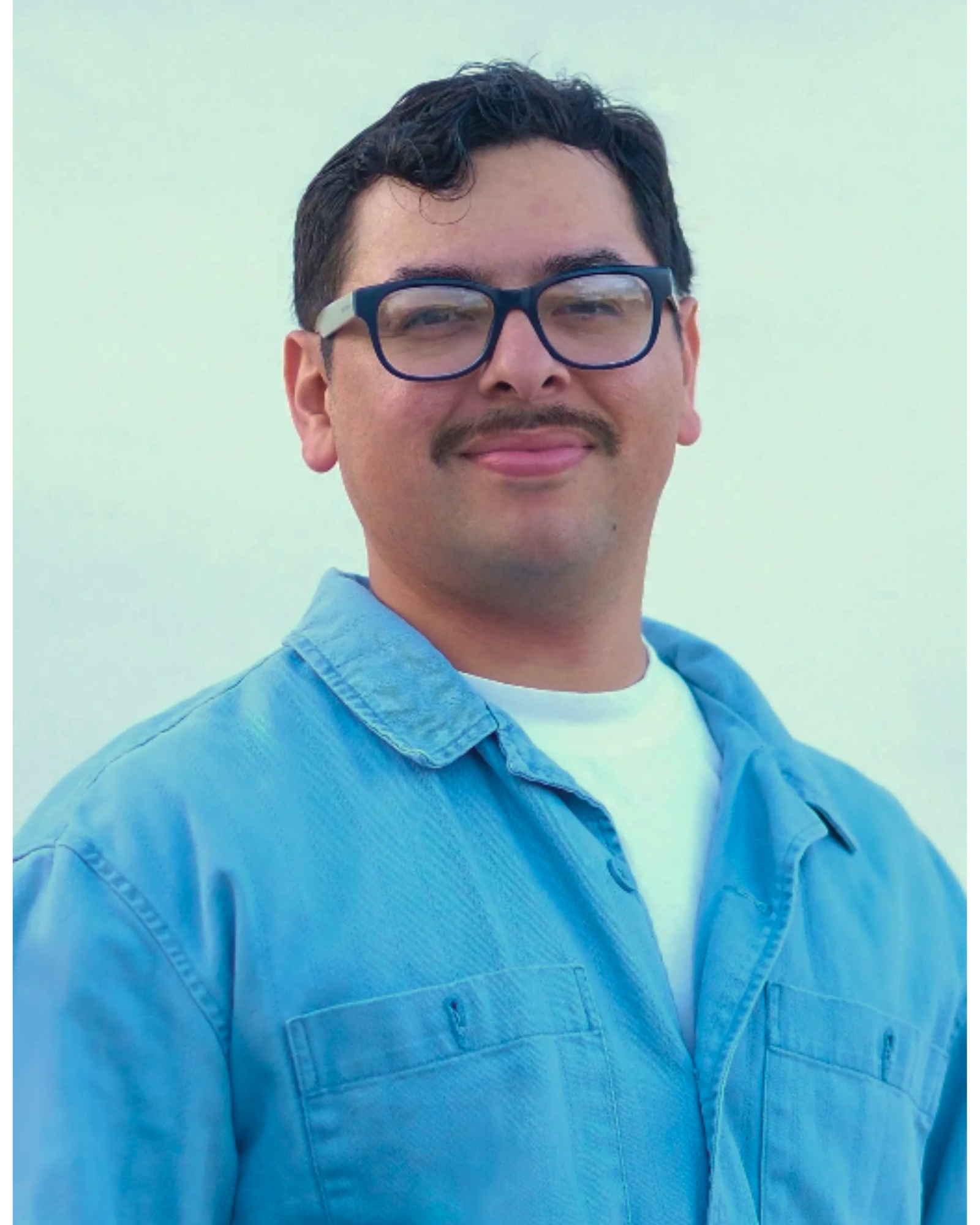 A man with dark hair, wearing glasses, a light blue jacket over a white shirt, standing outdoors with a plain sky background.