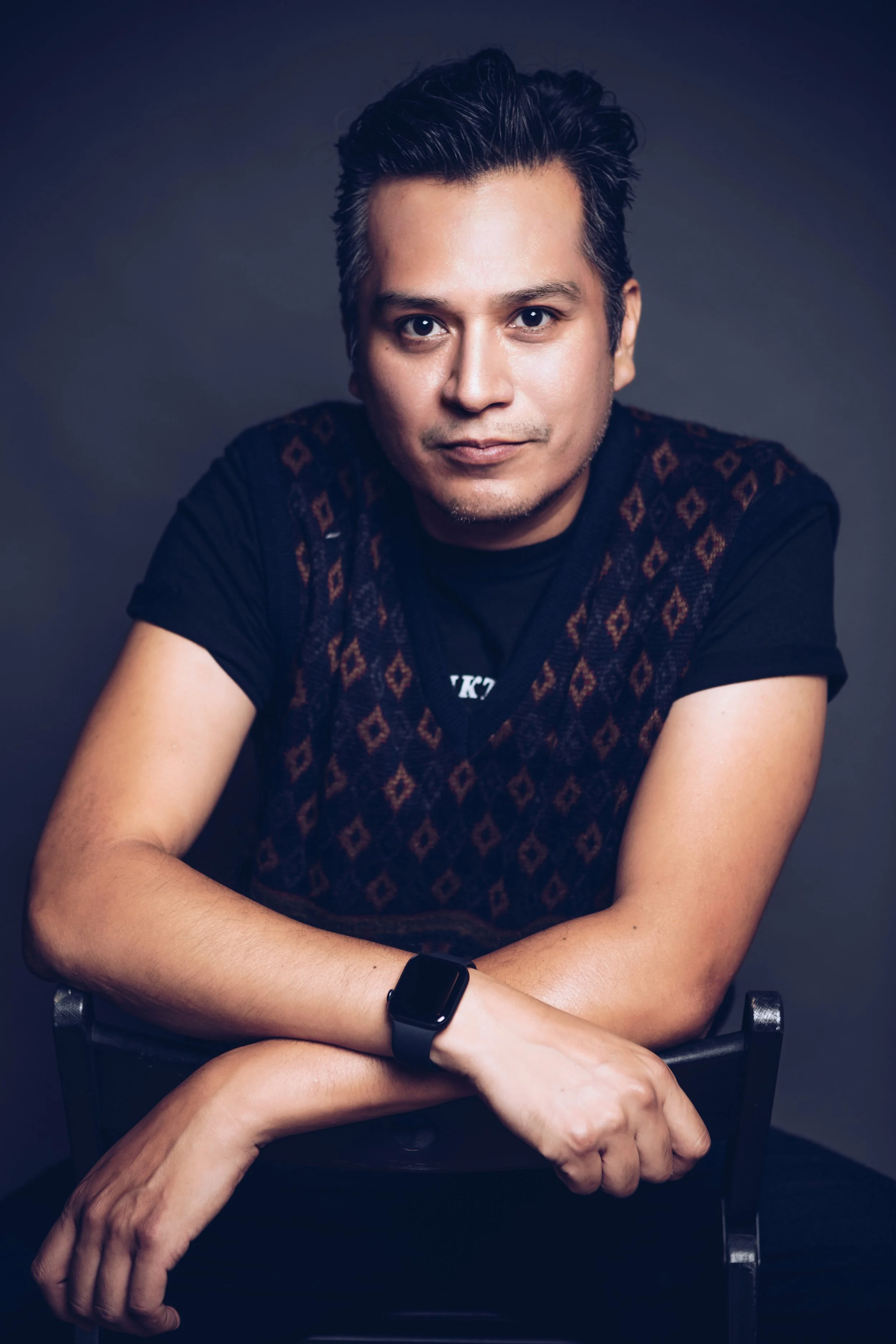 A man with dark hair styled back, wearing a black shirt and a sweater vest with a diamond pattern, sitting with crossed arms, looking at the camera, against a dark background.