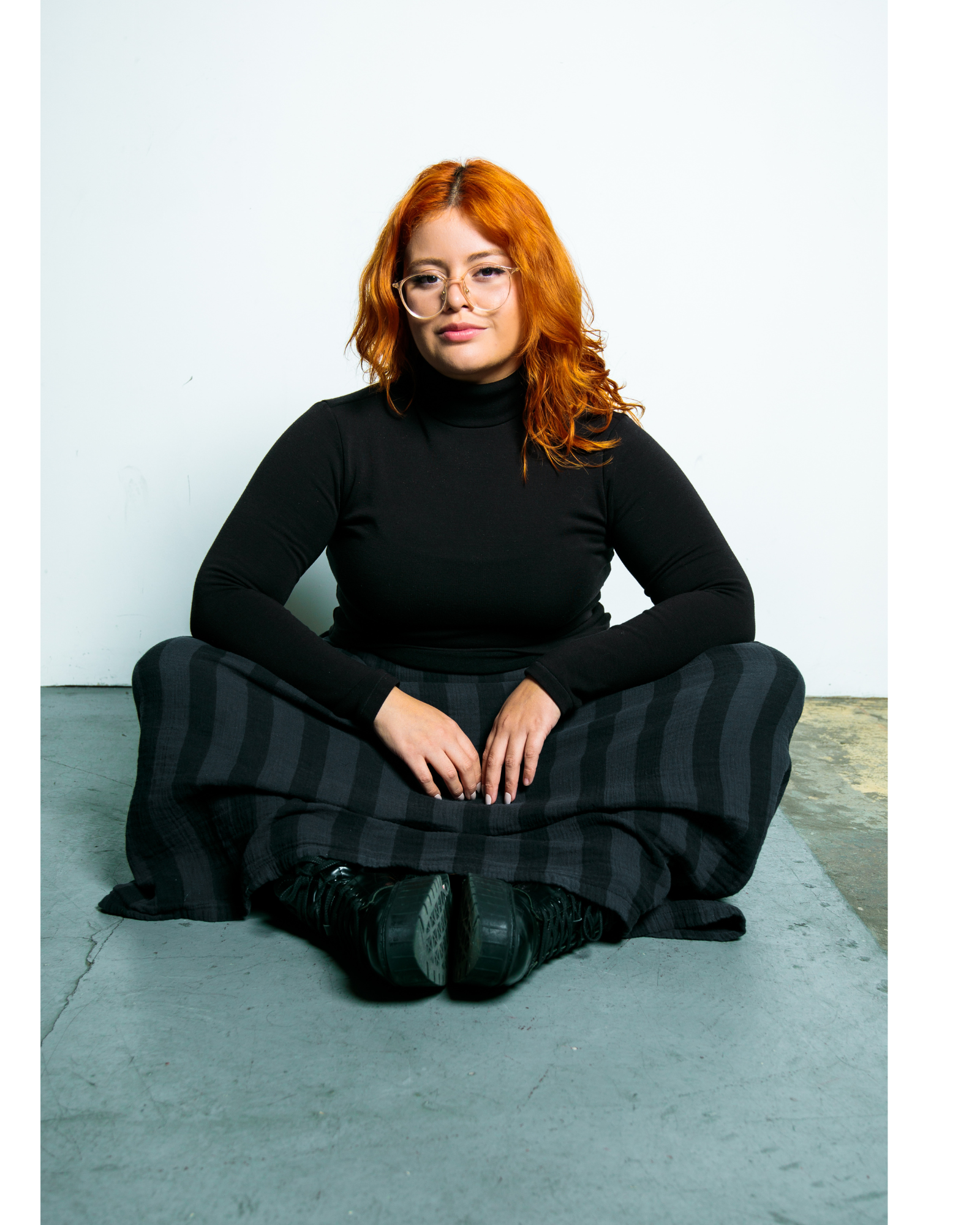 A woman with red hair, glasses, wearing a black turtleneck, striped wide-leg pants, and black shoes, sitting cross-legged on the floor against a white wall.