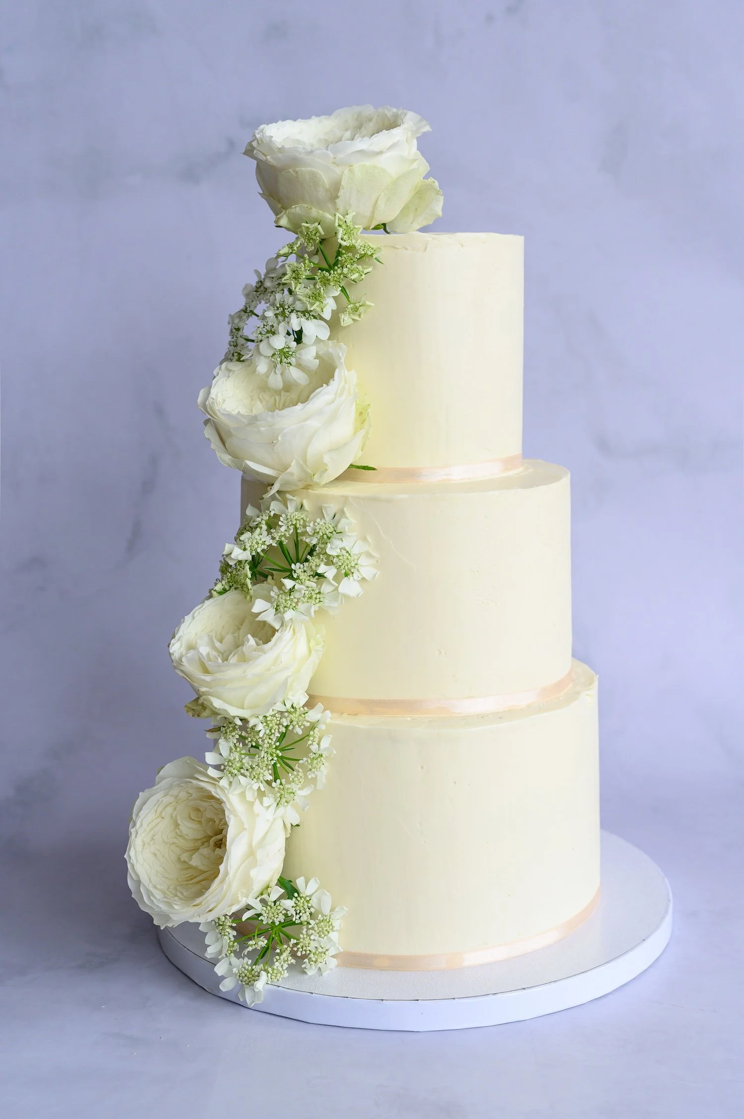 A four-tiered white wedding cake decorated with white roses and small white flowers, set against a light blue background.