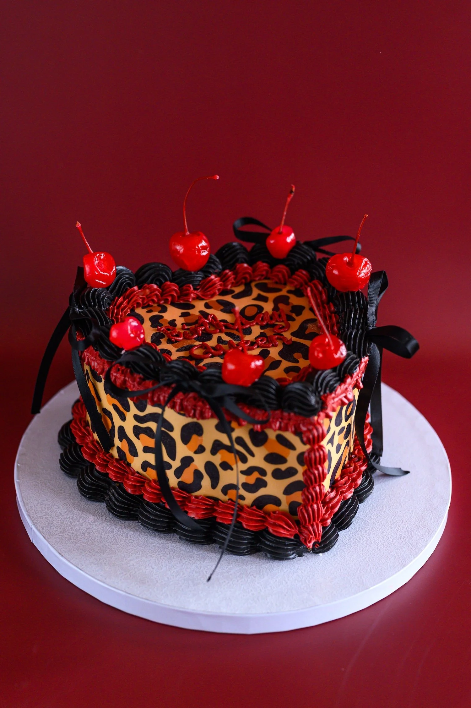 A square-shaped birthday cake decorated with leopard print fondant, black and red frosting piping, and topped with cherries, placed on a white round cake board.