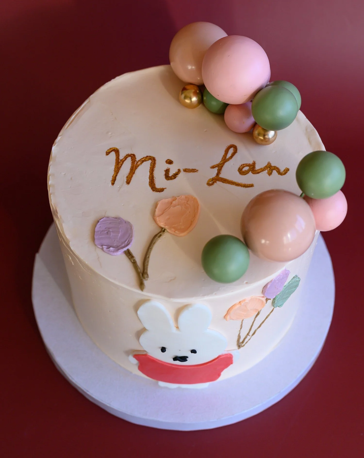 A birthday cake decorated with pastel-colored balloons, a bunny face, and the words 'Mi-Lan' written in gold.
