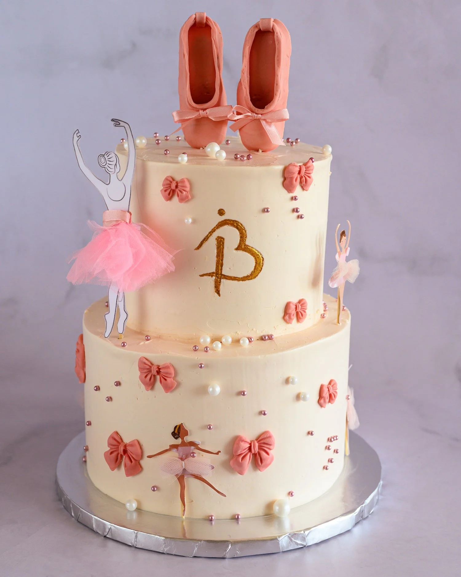 A three-tiered ballet-themed cake decorated with pink ballet shoes on top, pink bows, ballerina figurines, and edible pearls, with a light-colored background.