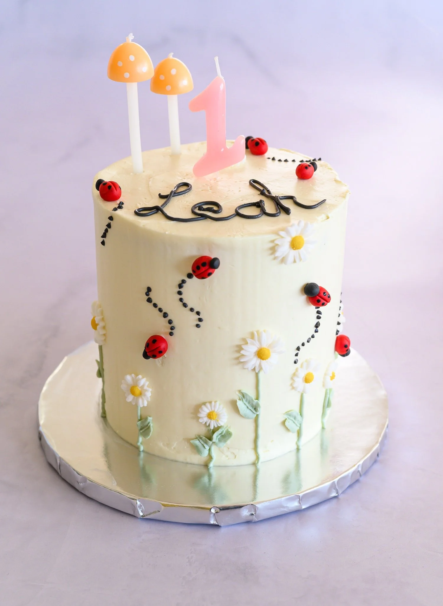White birthday cake decorated with daisies and ladybugs, topped with pink candle shaped like number one, and two orange mushroom-shaped candles.