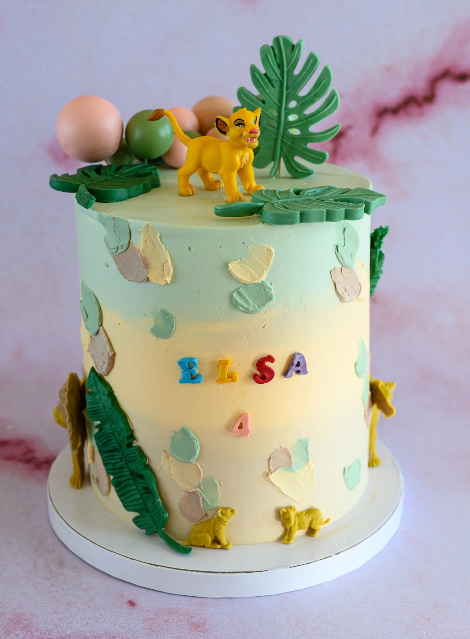 A decorated birthday cake with a lion figurine on top, tropical leaves, and pastel-colored decorations, with the word 'ELSA' on the side.