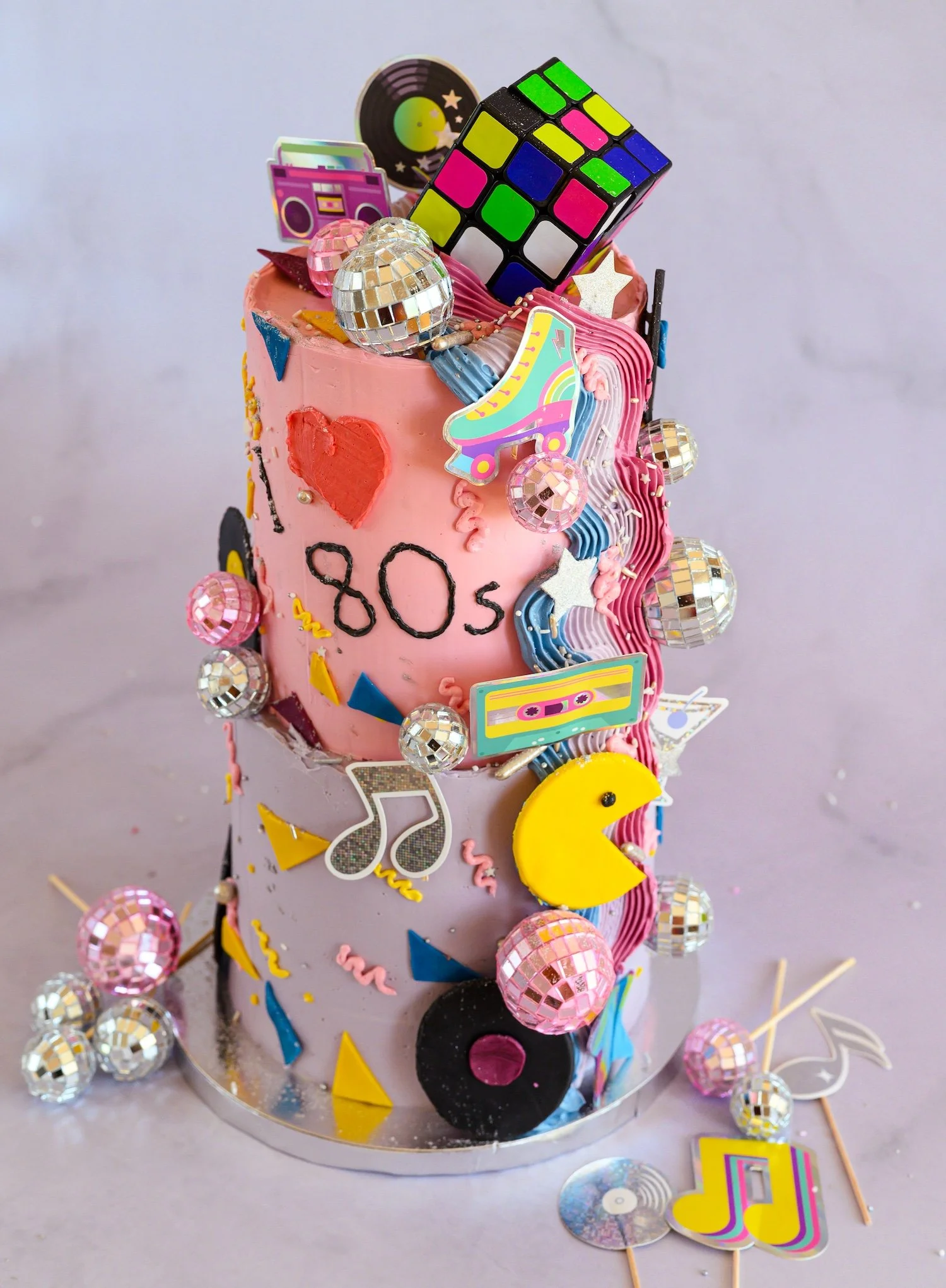 A birthday cake decorated with colorful pop-culture themed edible and non-edible decorations, including a Rubik's cube, disco balls, stickers, and small cupcake toppers.