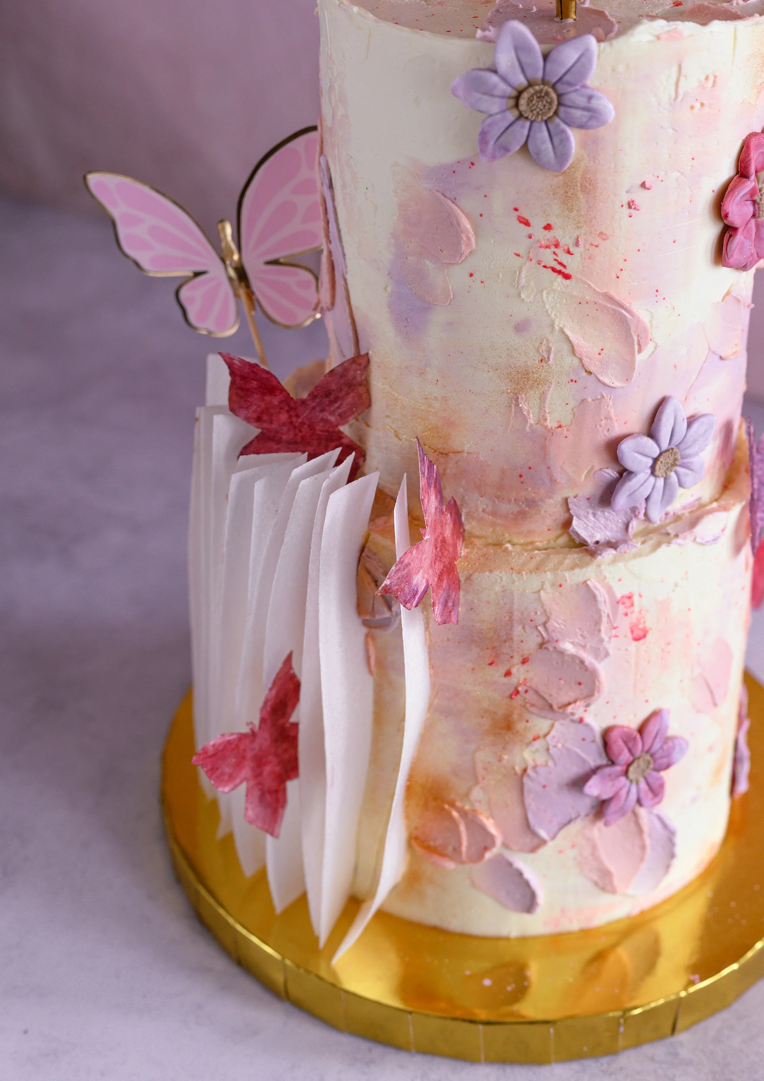 Decorated multi-tier pink cake with sugar flowers, butterflies, and feathers on a gold cake board.