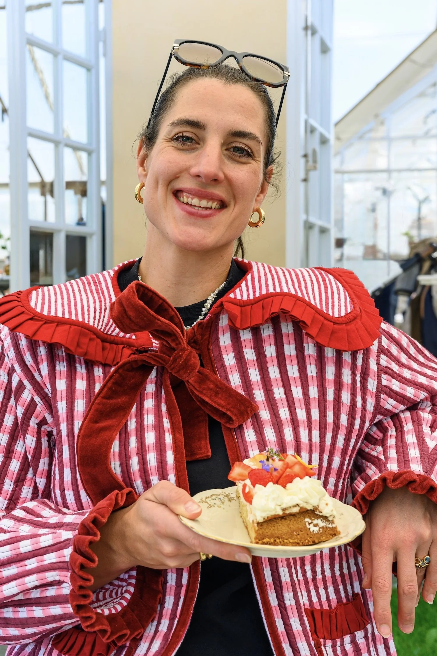 A woman wearing a red and white striped jacket with ruffled edges and golden hoop earrings, standing inside a greenhouse with large windows, holding a plate of cake topped with strawberries and cream, smiling at the camera.