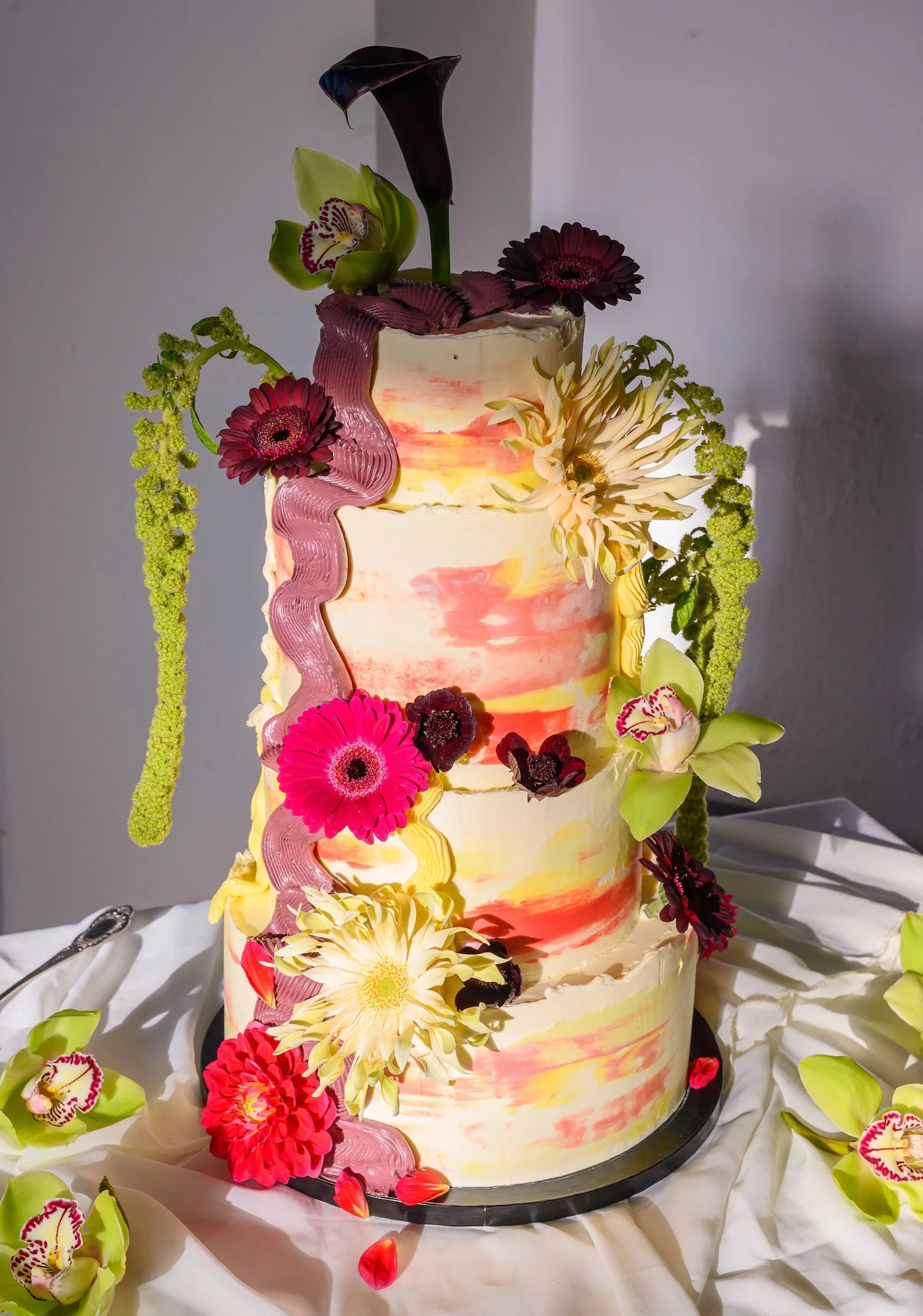 A multi-tiered wedding cake decorated with vibrant flowers, including gerbera daisies, orchids, and calla lilies, topped with dark purple Calla lily flowers.