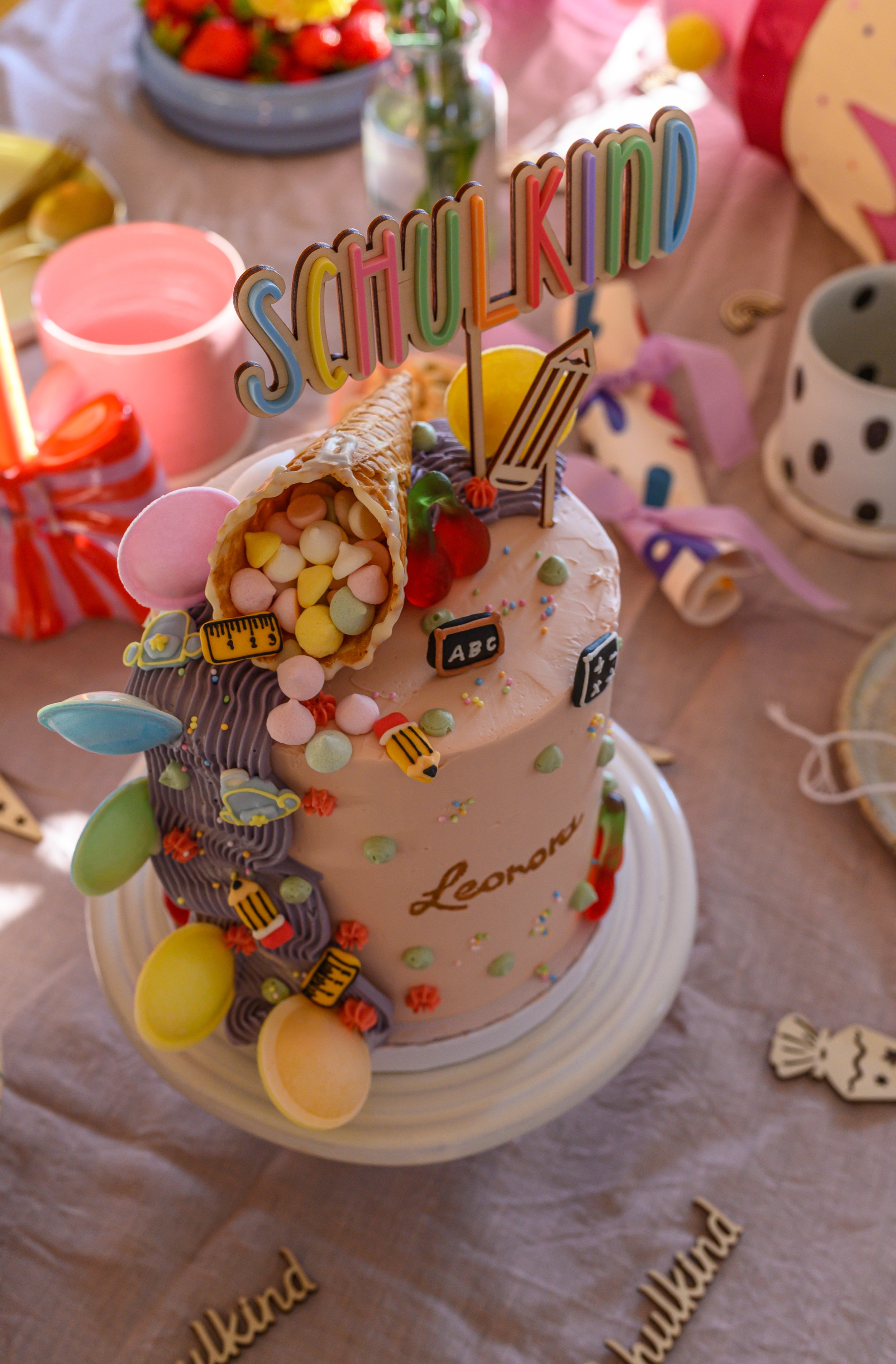 Colorful birthday cake with the word 'SQUILD KID' on top, decorated with candies, a rolled wafer, and cupcake toppers, on a table with other party decorations and snacks in the background.
