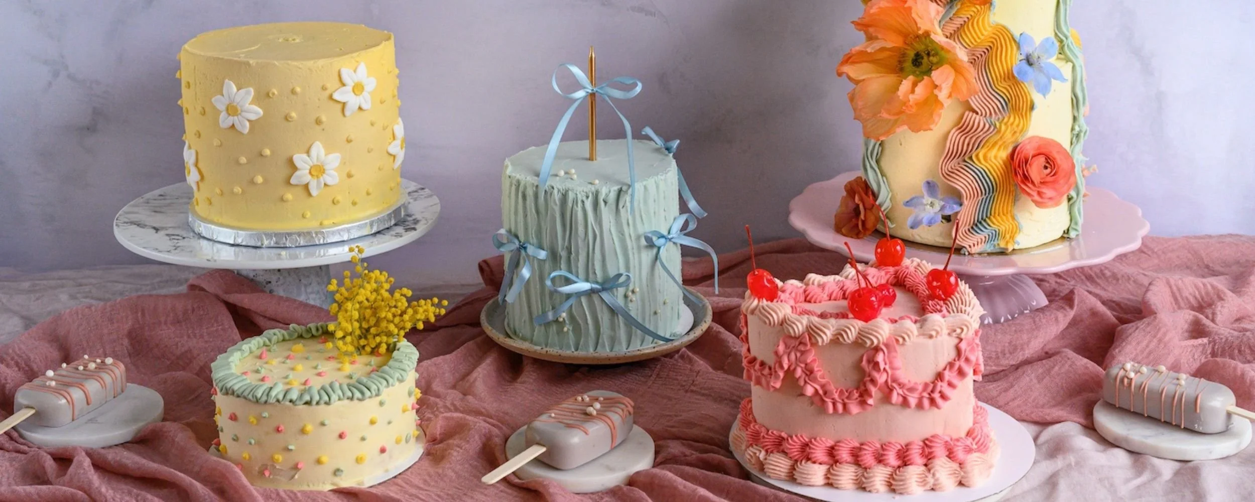 Assorted decorated cakes and popsicles on a table with pink fabric