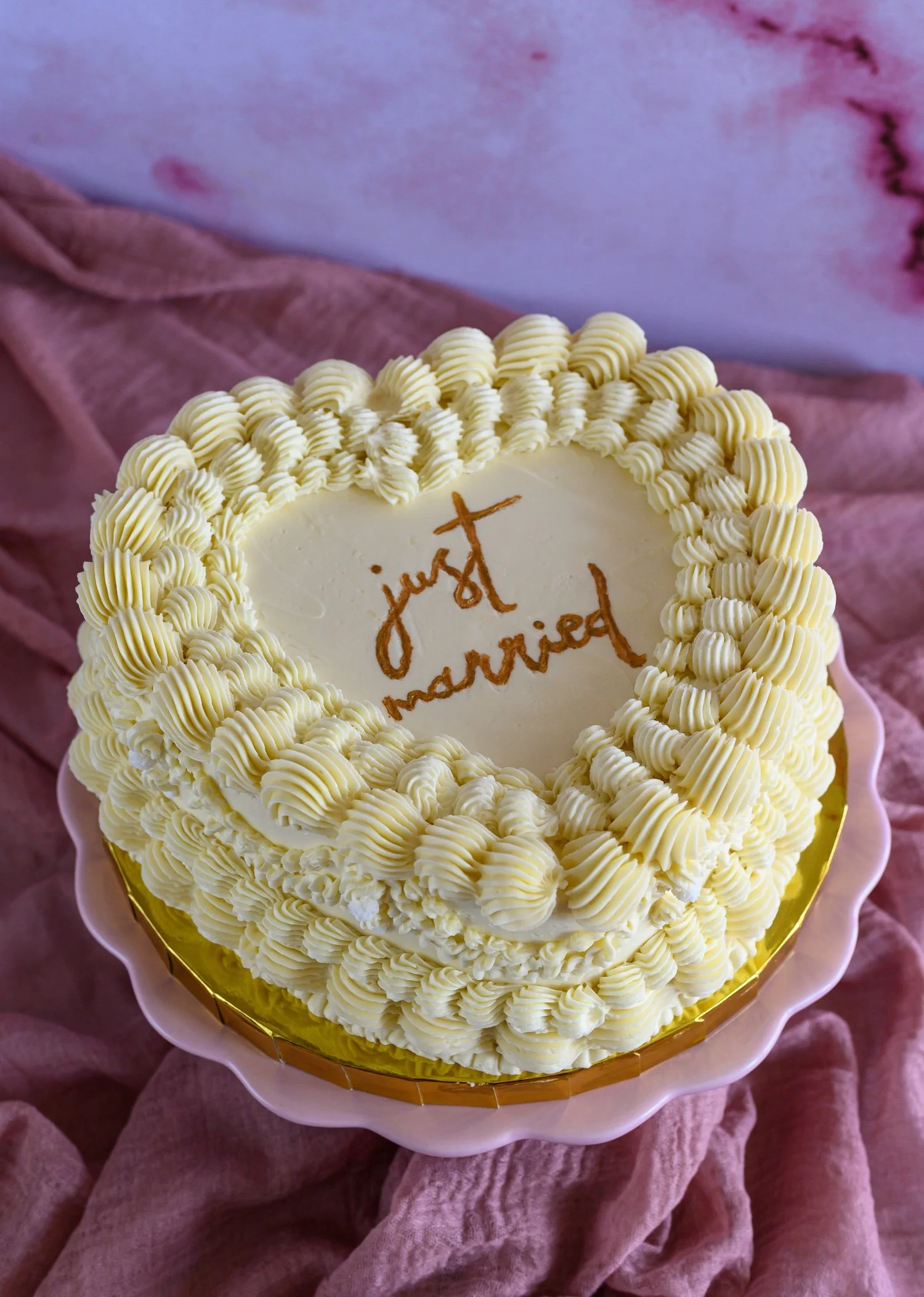 A round white cake with decorative piped cream borders on top and around the edge, with a message written in brown on the top that says "Just Married." The cake is placed on a pink, ruffled fabric surface.