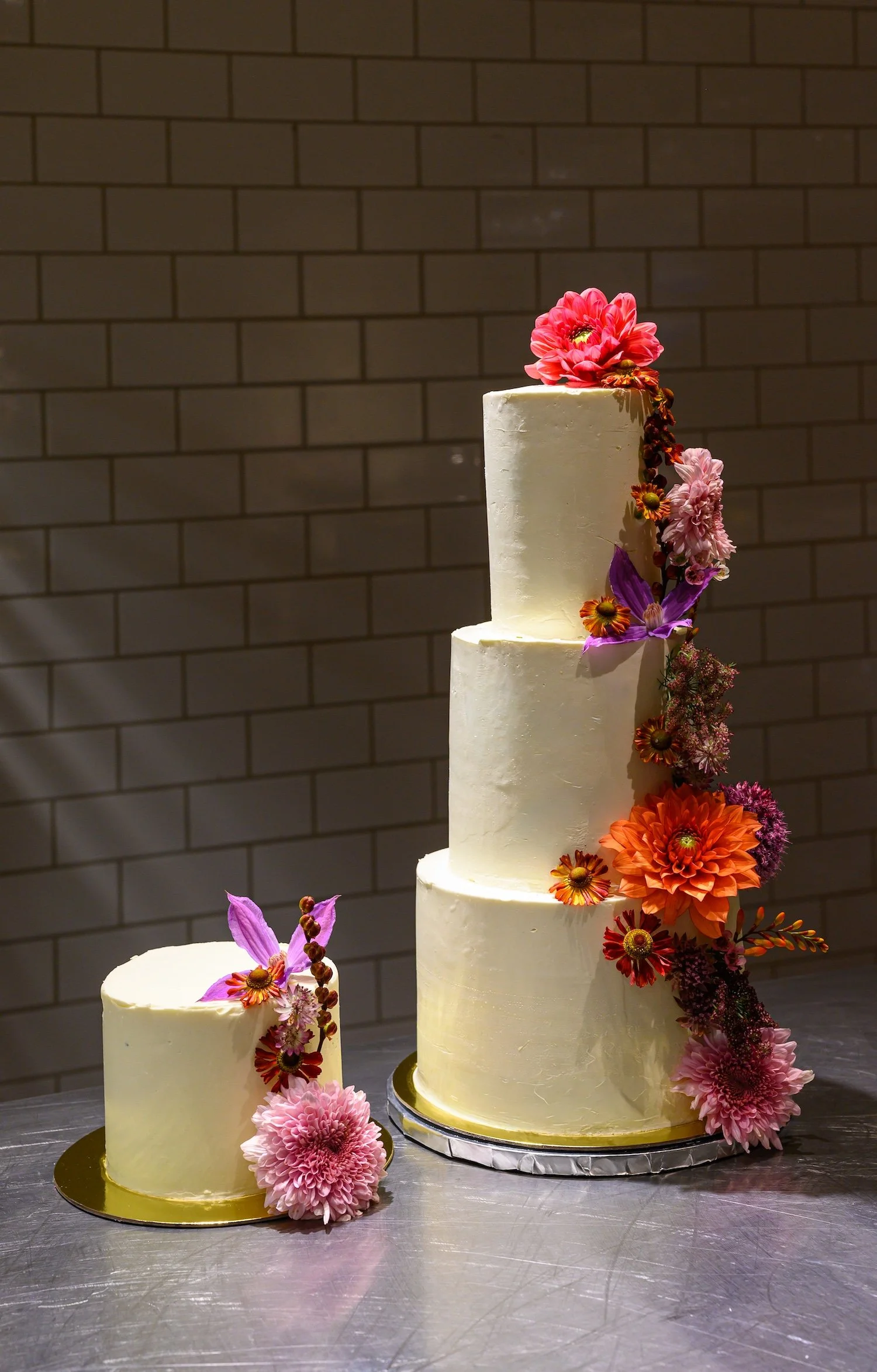 A three-tier white wedding cake decorated with pink and orange flowers, including dahlias and lilies, set against a brick wall background.