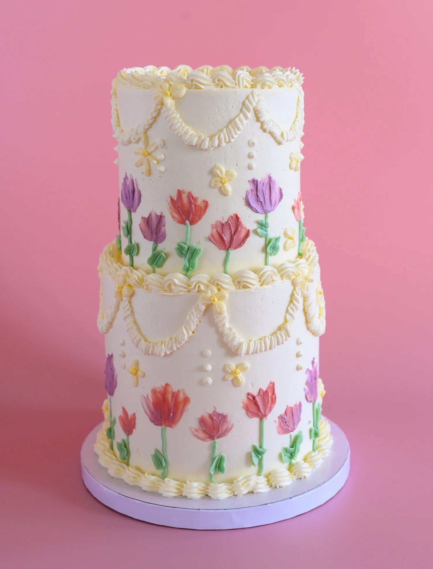 A tall, white, multi-layered birthday cake decorated with colorful flowers, pearls, and piped borders, set against a pink background.