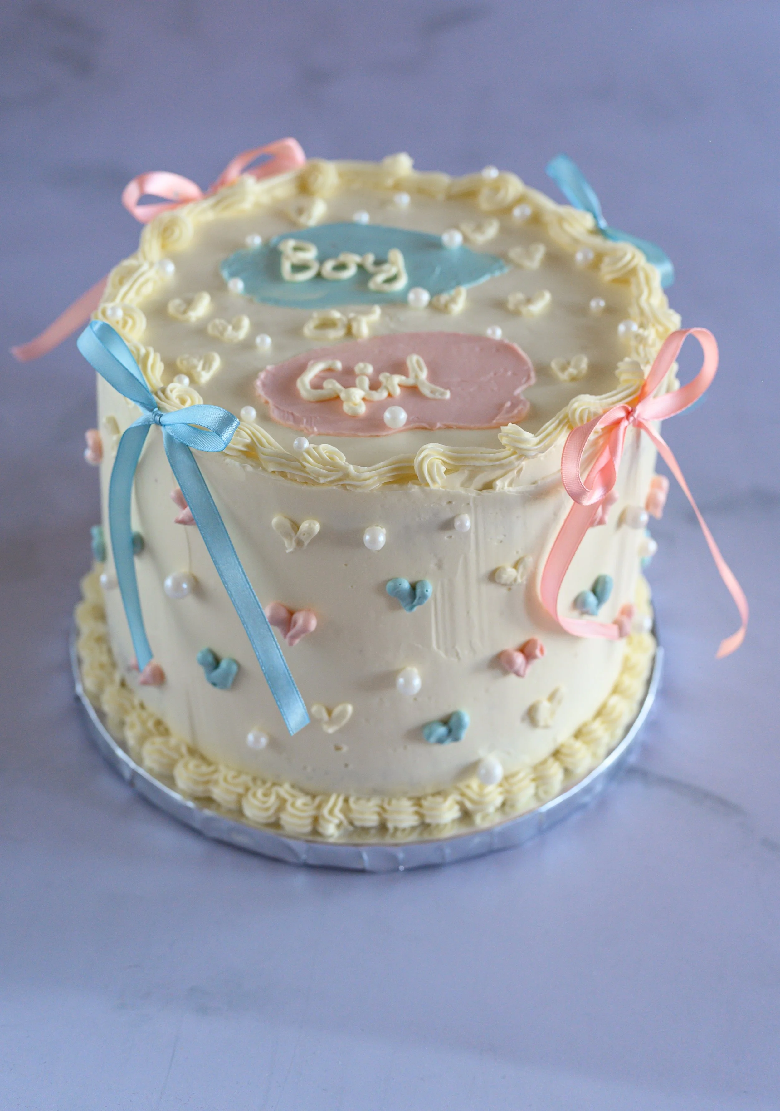 A white frosted cake decorated with pastel ribbons, small white pearls, and tiny heart-shaped decorations. The top of the cake has pink and blue accents with the words 'Boy' and 'Girl' written on different colored fondant shapes.