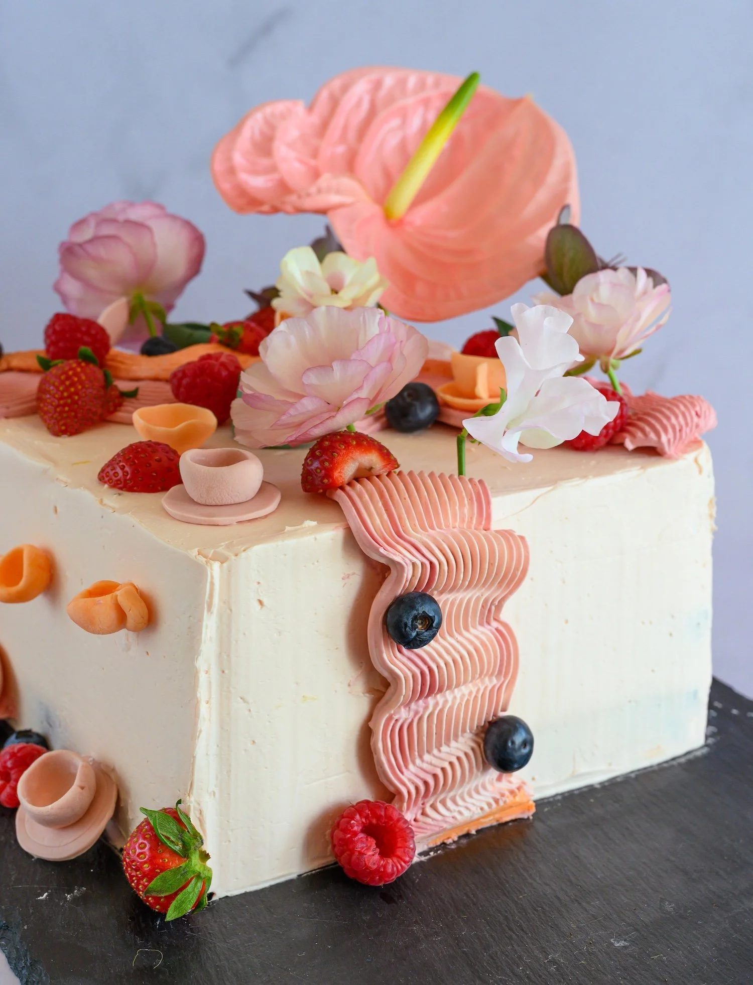 A decorated cake with strawberries, raspberries, blueberries, and flowers on top, featuring pink, peach, and white icing accents.