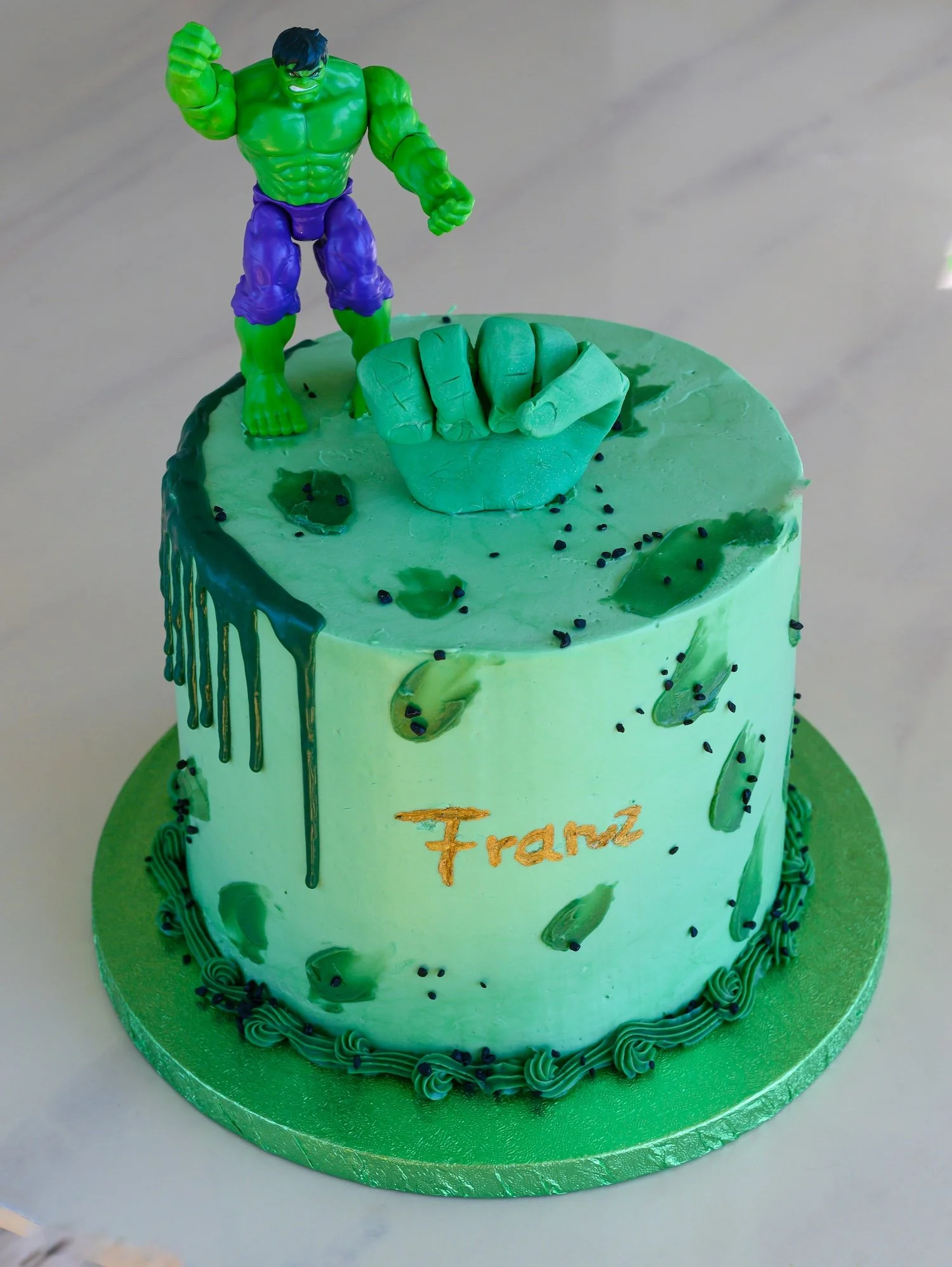 A green birthday cake decorated with a Hulk figure on top, holding a large green fist. The cake has green and black sprinkles, green icing drips, and the name 'Frank' written in gold on the front.
