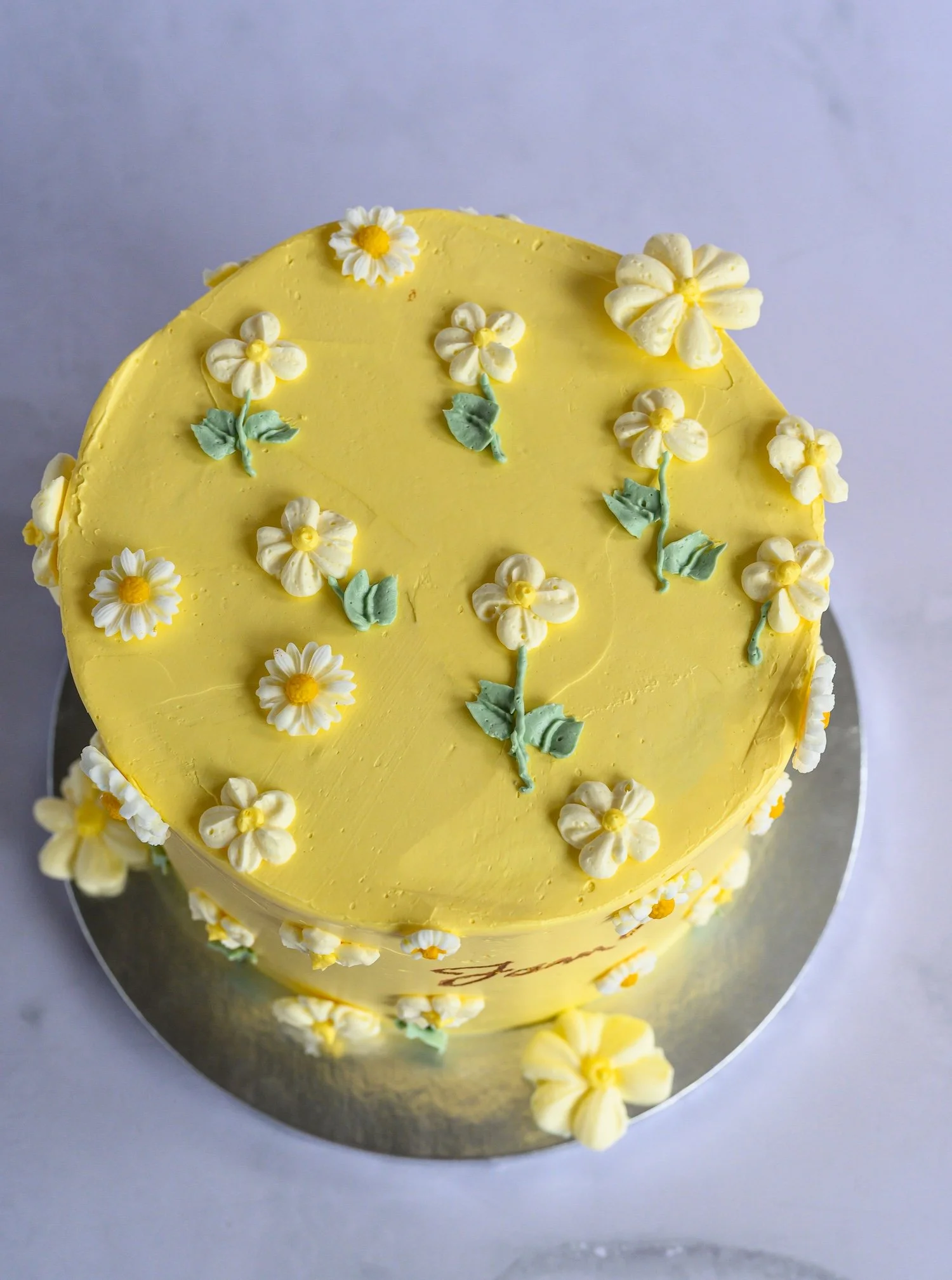 Yellow round cake decorated with white and yellow flower-shaped frosting and green leafy accents.