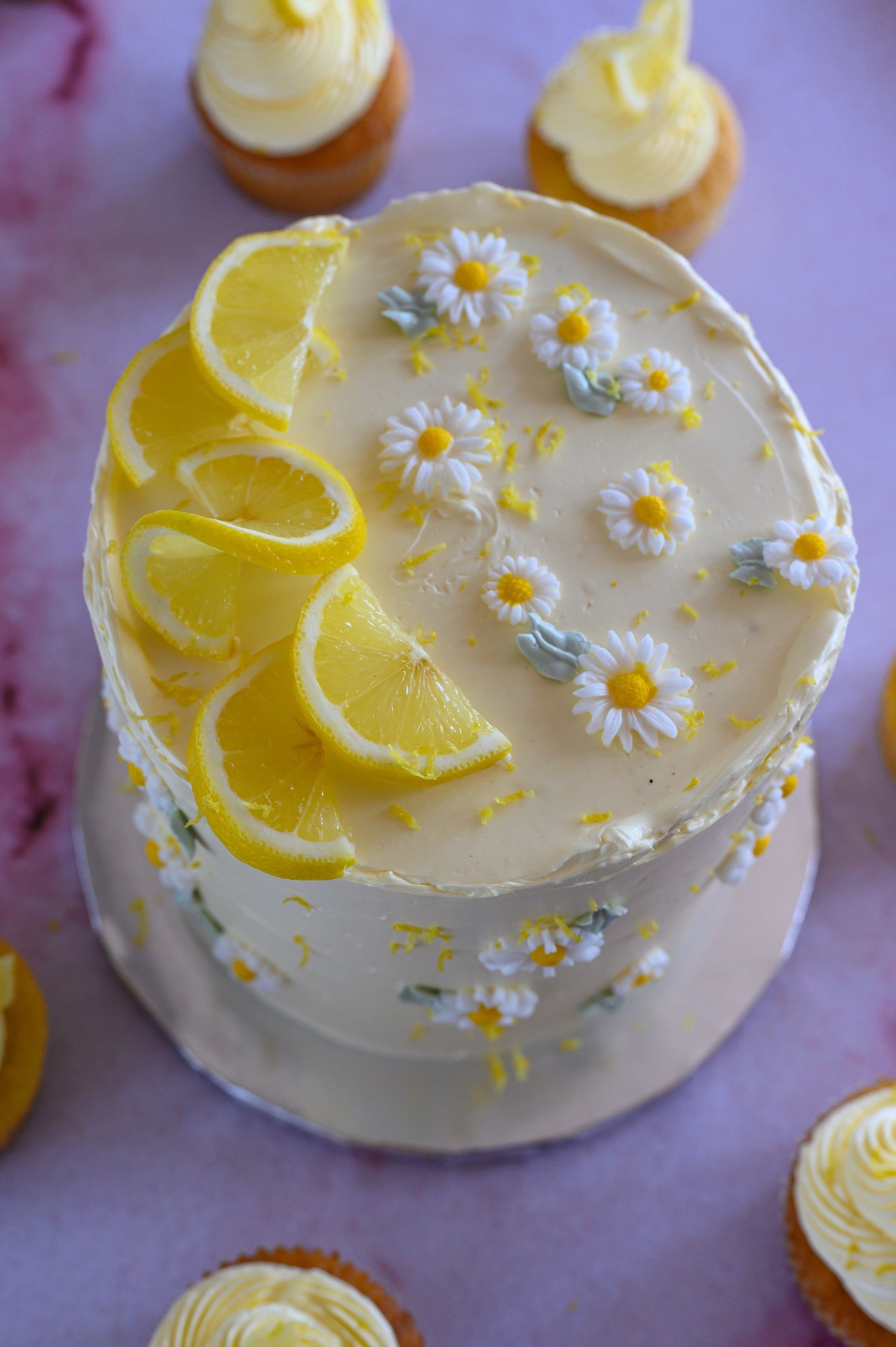 A round lemon cake decorated with lemon slices and white daisies with yellow centers and green leaves, topped with white icing.