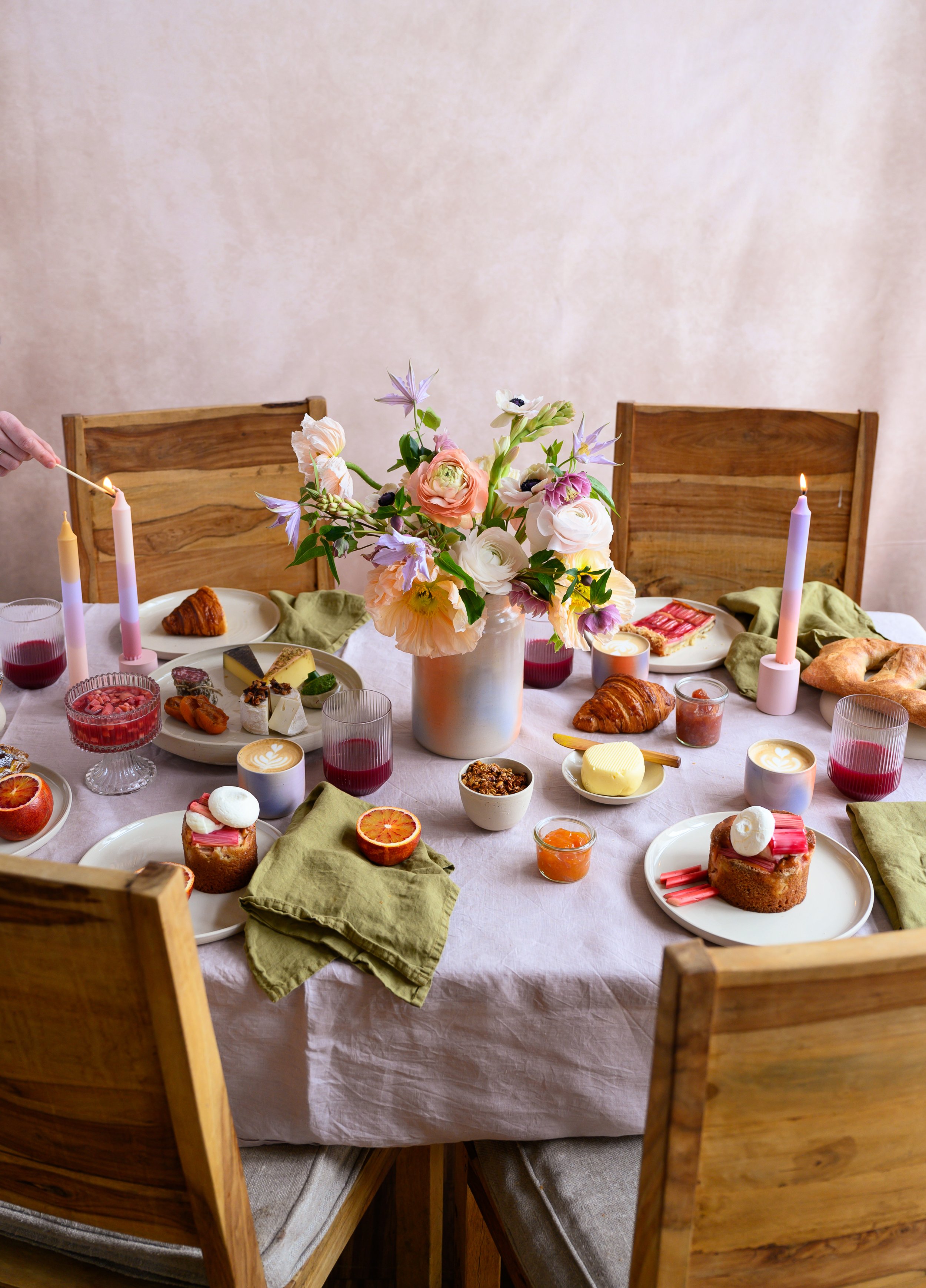 A festive dining table set with a pink tablecloth, green napkins, and a large centerpiece of pink and white flowers in a white vase. The table features an assortment of desserts, including cakes, croissants, and fruit, with candles and glasses of red wine.