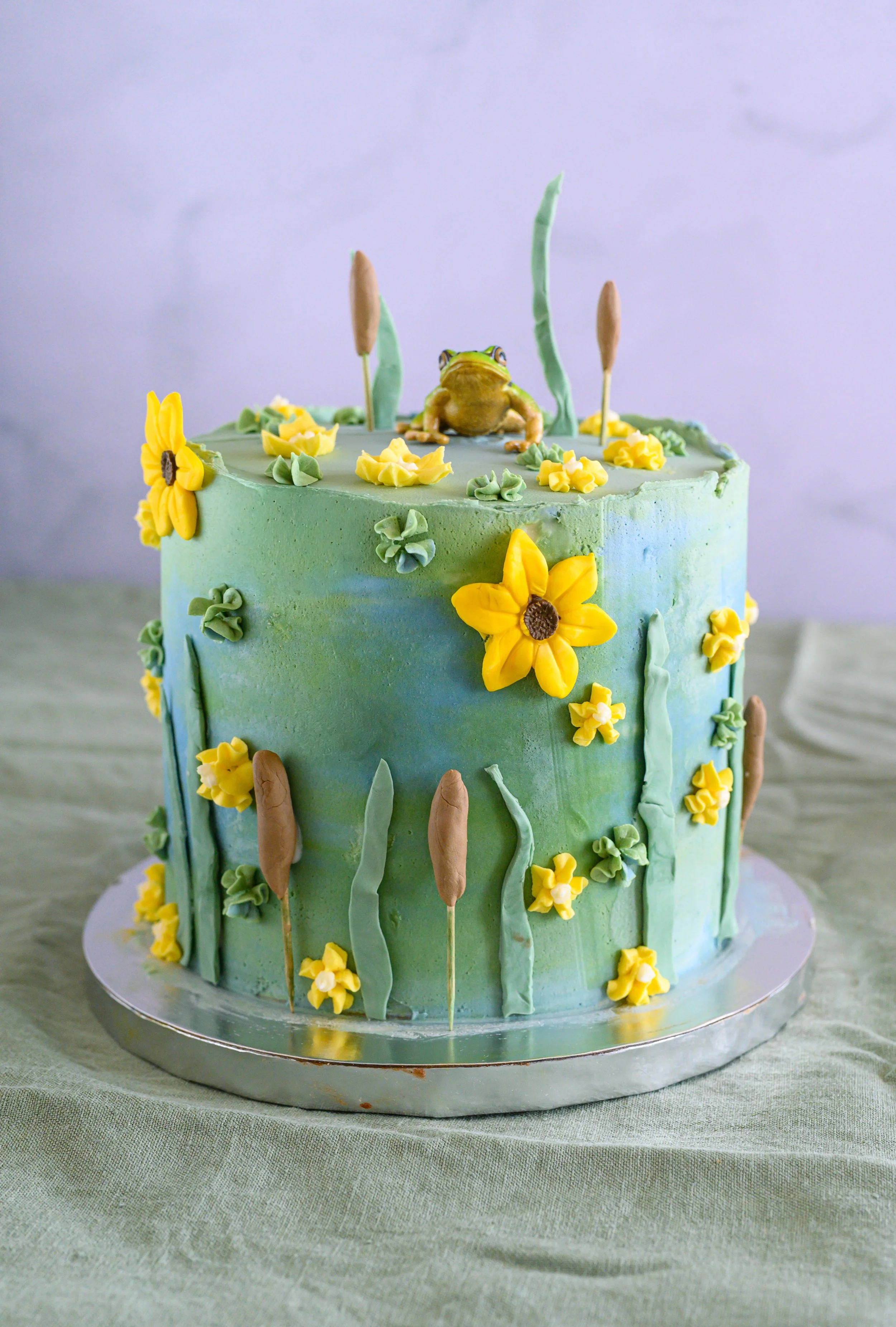A two-tiered cake decorated with yellow and green flowers, green grass-like piping, and a frog figurine on top, set on a silver cake board.