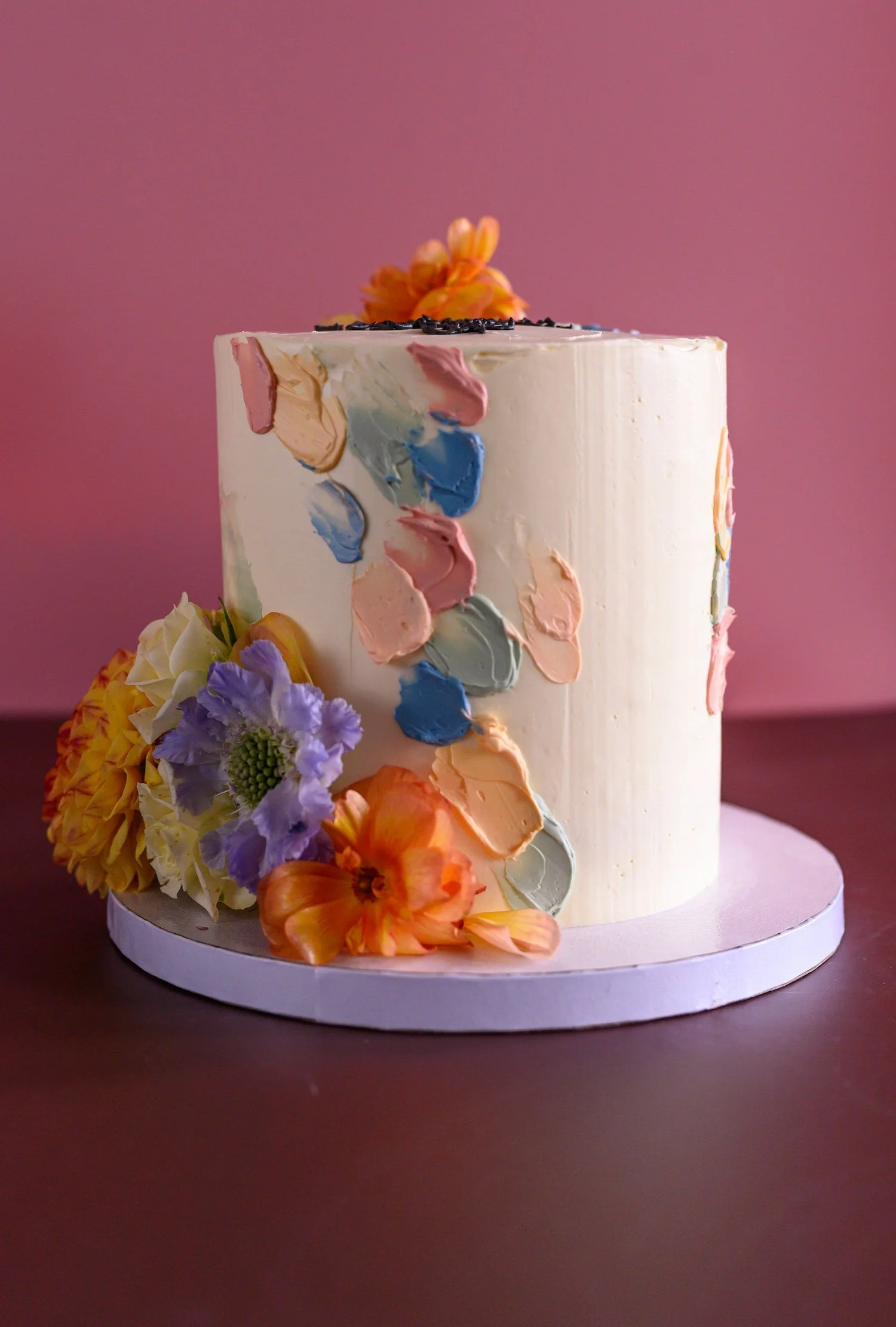 A tall white cake decorated with colorful flower and petal icing, topped with a yellow flower and black sprinkles, placed on a white cake stand, with lilac, peach, and yellow flowers at the base and a pink background.