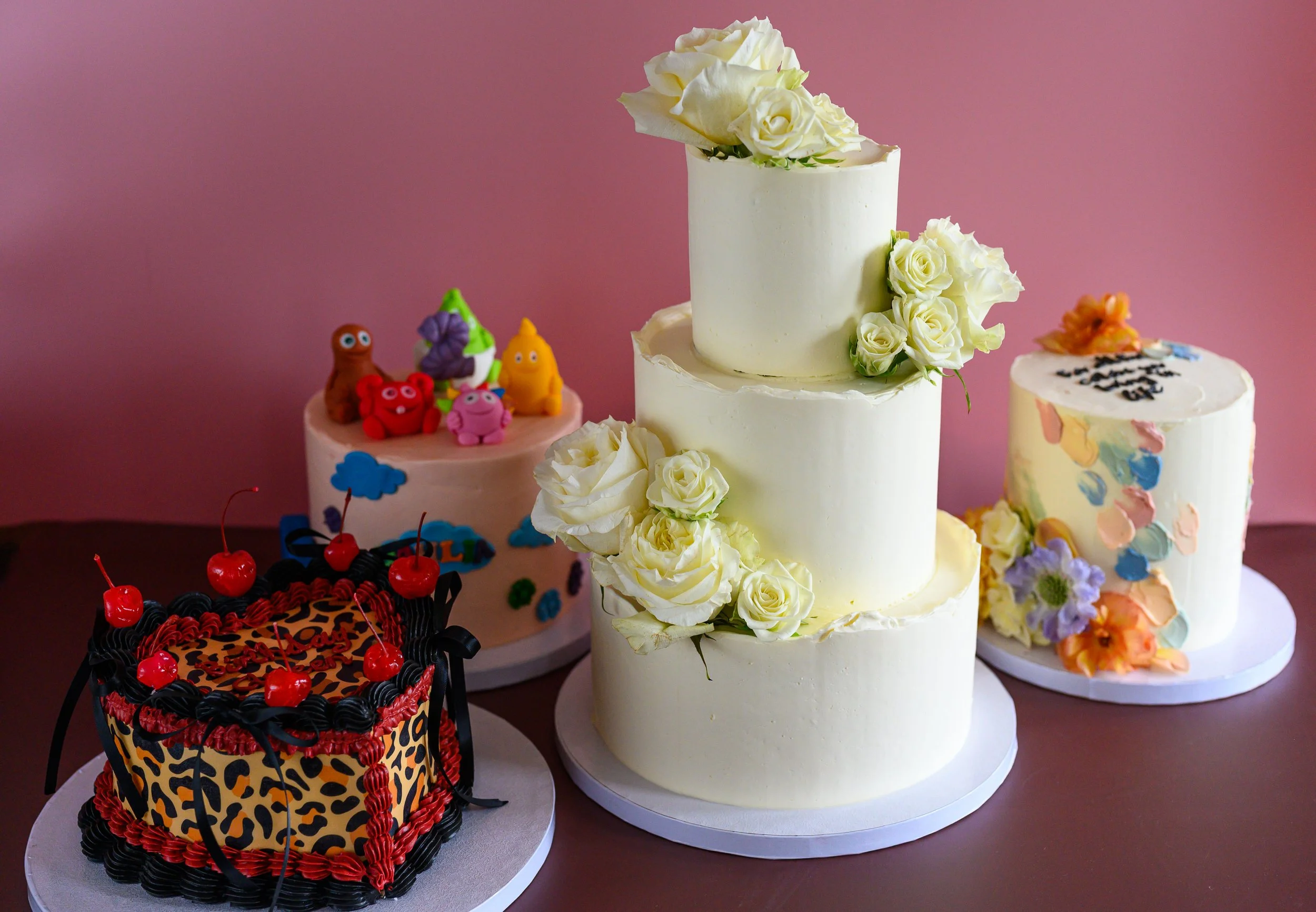 Collection of decorated birthday cakes on a table against a pink background, featuring various designs including chocolate with cherries, floral, and cartoon character themes.