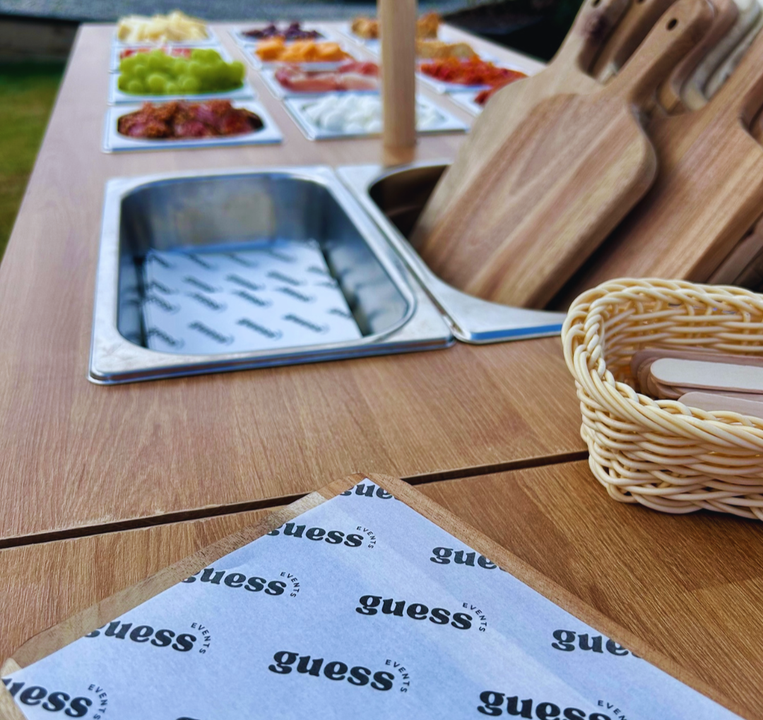 A wooden table set up outdoors with various food items on plates, a metal serving tray, wooden cutting boards, and a basket of utensils. The background shows a grassy area.