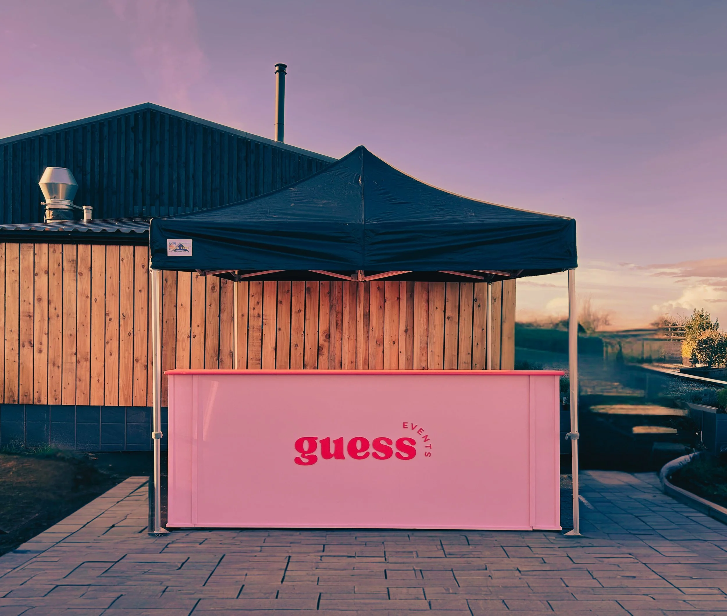 A pink event table with the Guess logo in front of a wooden and blue building. There is a black canopy above the table and a paved walkway in front.