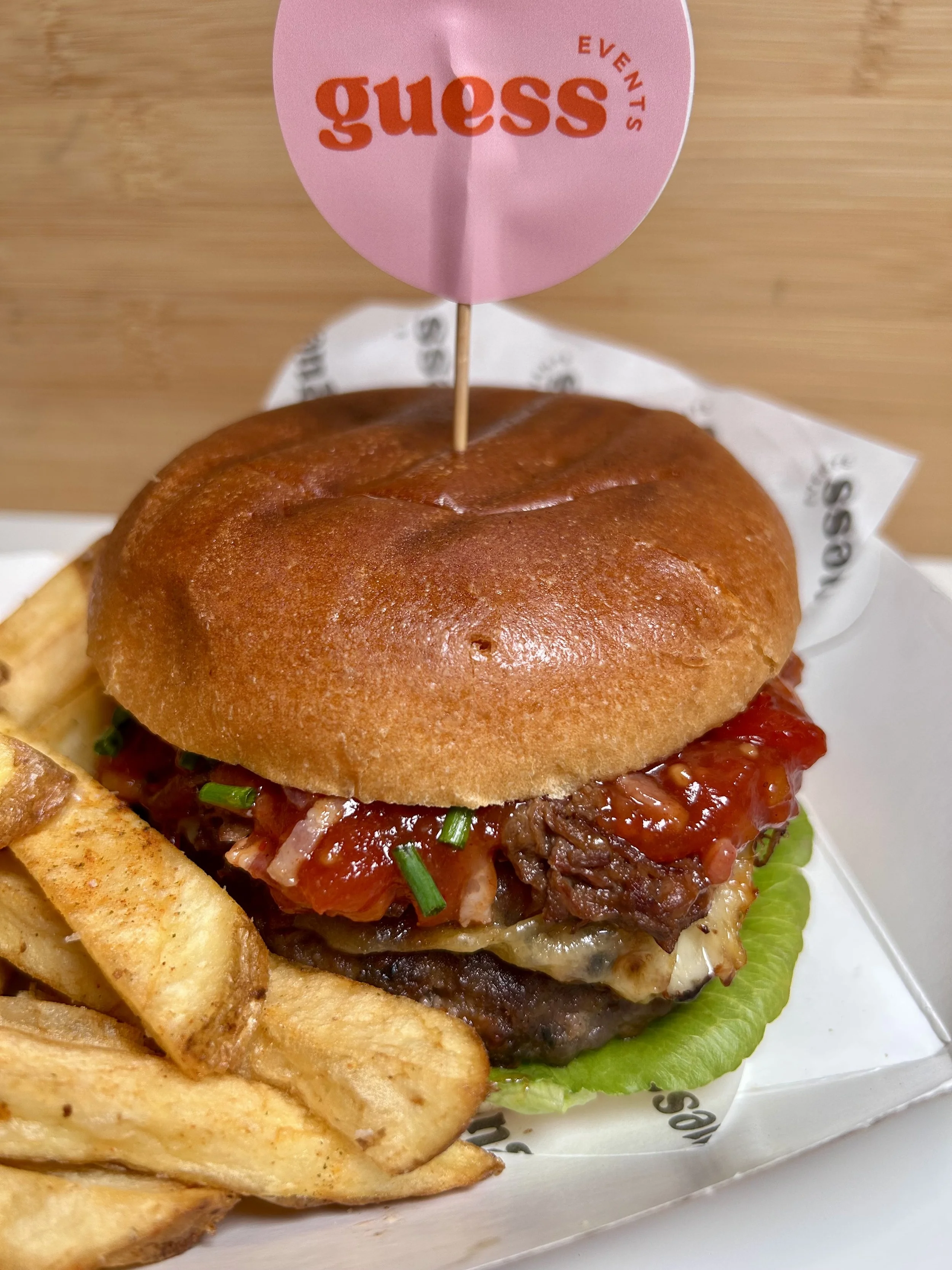 Close-up of a hamburger with lettuce, cheese, beef patty, tomato, bacon, topped with barbecue sauce, in a bun, with a side of seasoned French fries, and a pink flag with 'Guess Events' on it.