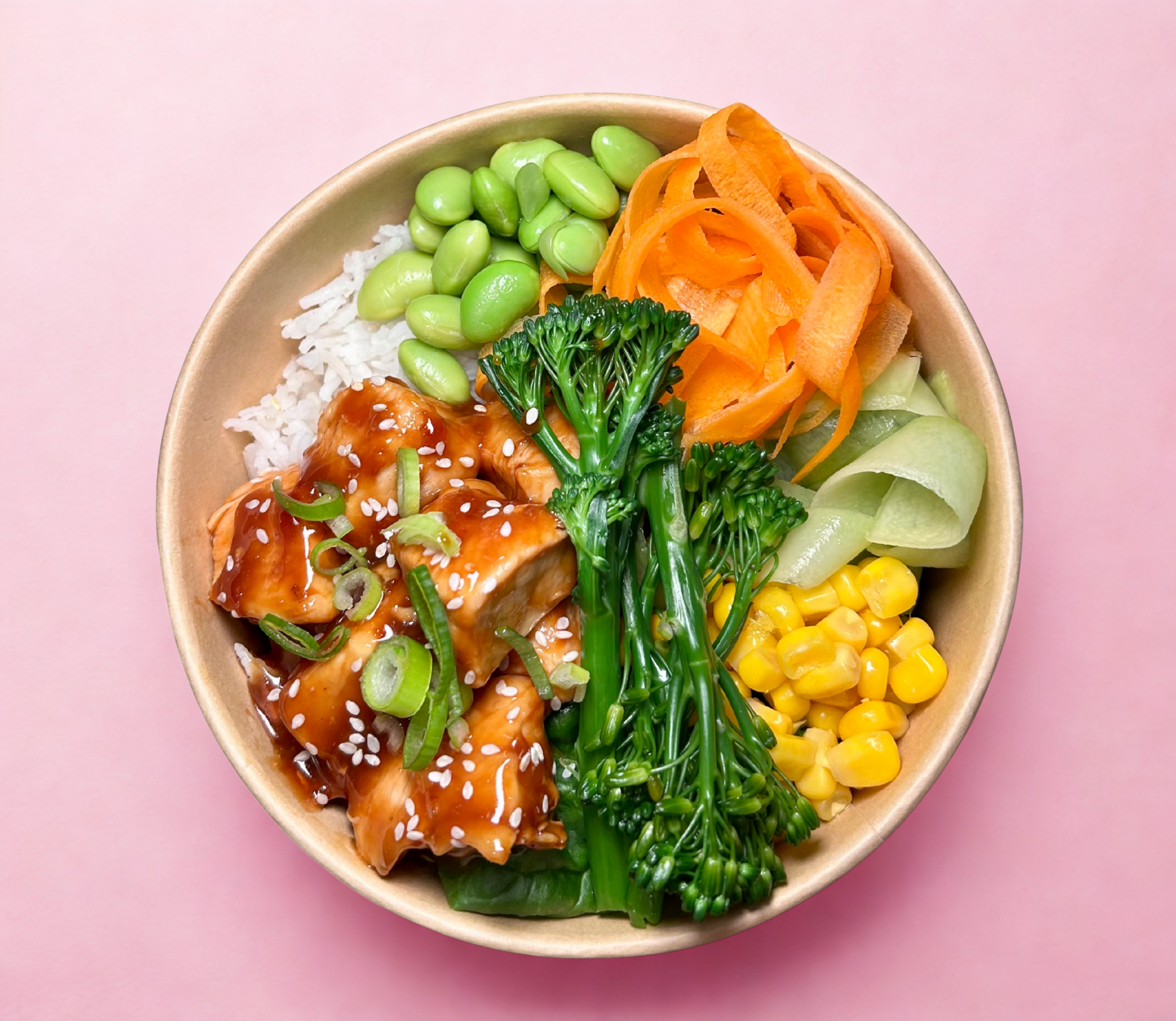 A bowl of assorted vegetables and rice, including edamame, shredded carrots, thin cucumber slices, broccoli, corn, green onions, and pieces of cooked chicken topped with sesame seeds on a pink background.