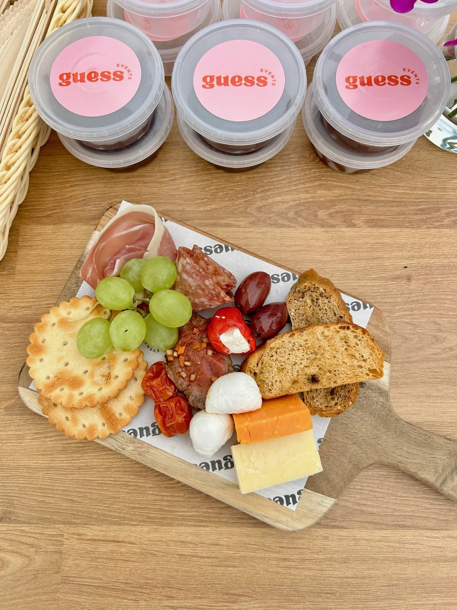 A cheese and meat platter with crackers, grapes, cherry tomatoes, and cheese on a wooden serving board, with three containers labeled 'guess events' on the table in the background.