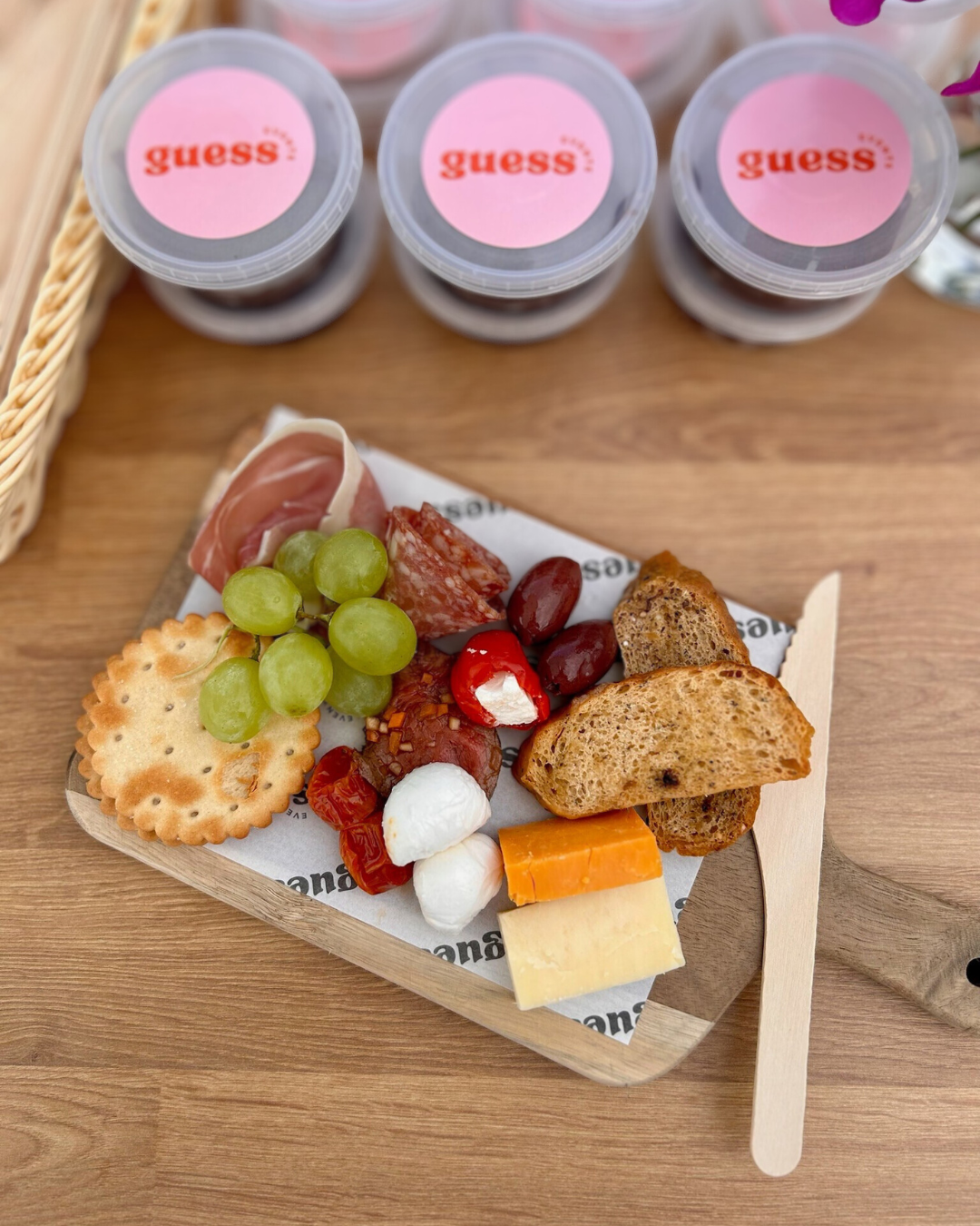 A cheese and charcuterie board with grapes, crackers, cured meats, olives, cherry tomatoes, mozzarella, and bread, set on a wooden tray with a wooden fork. In the background, there are three containers of dessert with pink labels that read "guess."