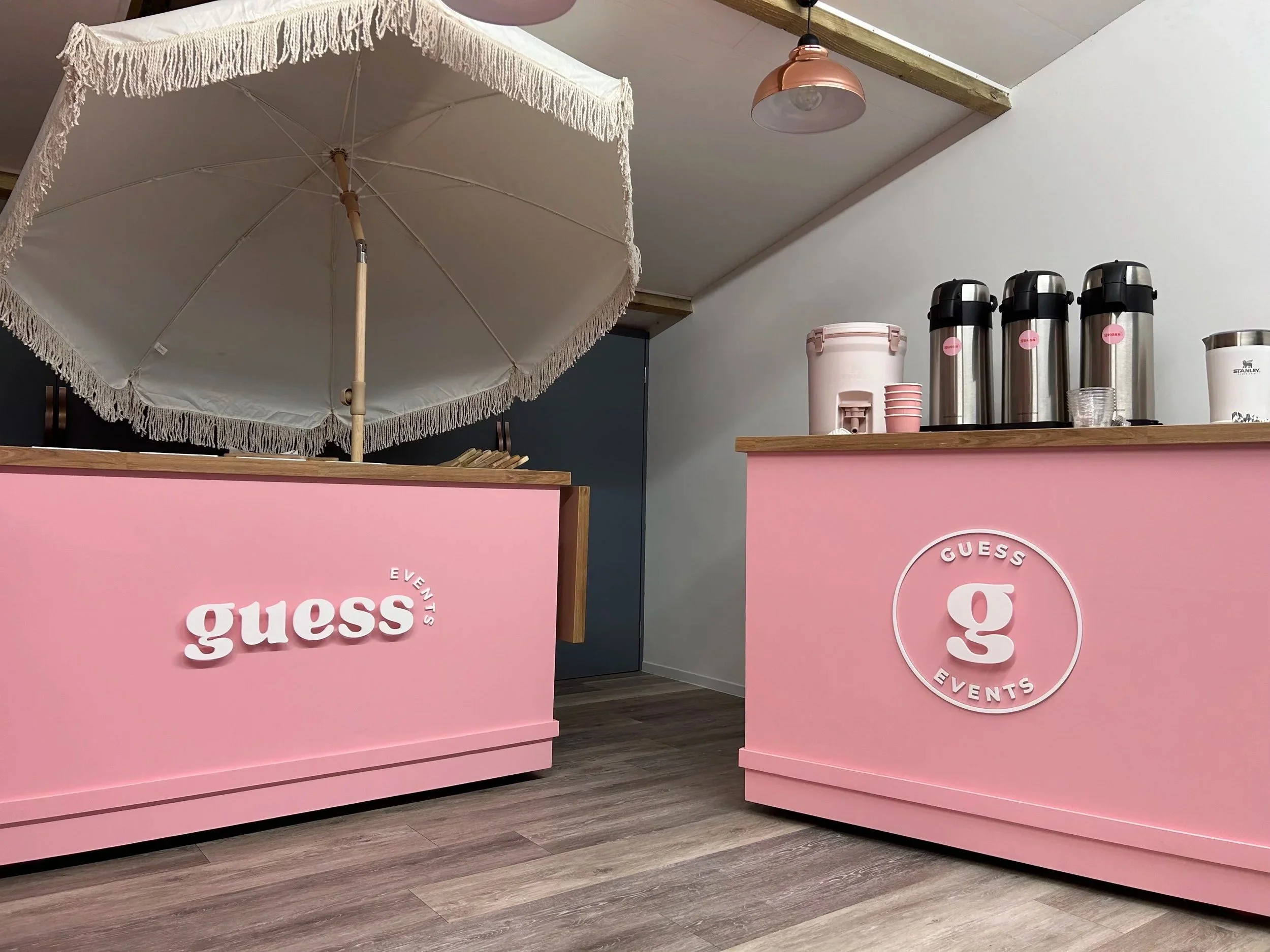 Pink counter with 'Guess Events' logo, white umbrella, and beverage dispensers on the counter.