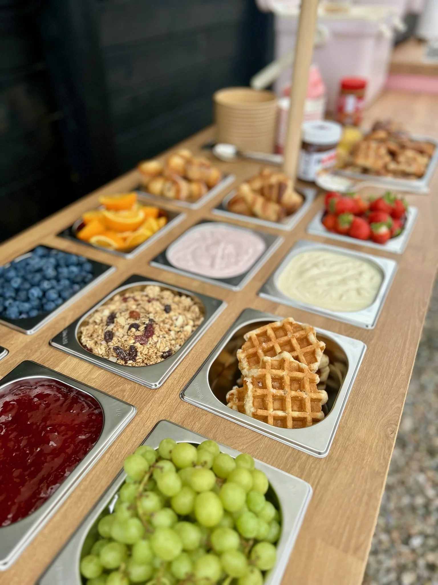 A buffet table with various breakfast foods including grapes, waffles, granola, blueberries, strawberries, sliced oranges, and containers of yogurt or cream.