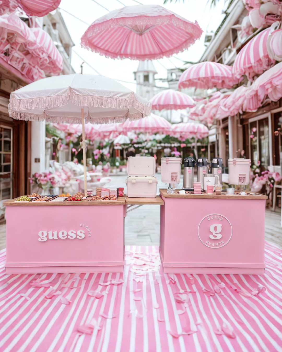 Pink themed outdoor event setup with two pink counters, umbrellas, and tablecloths, featuring beverages and snacks, branded with 'Guess' for a Guess Events occasion.
