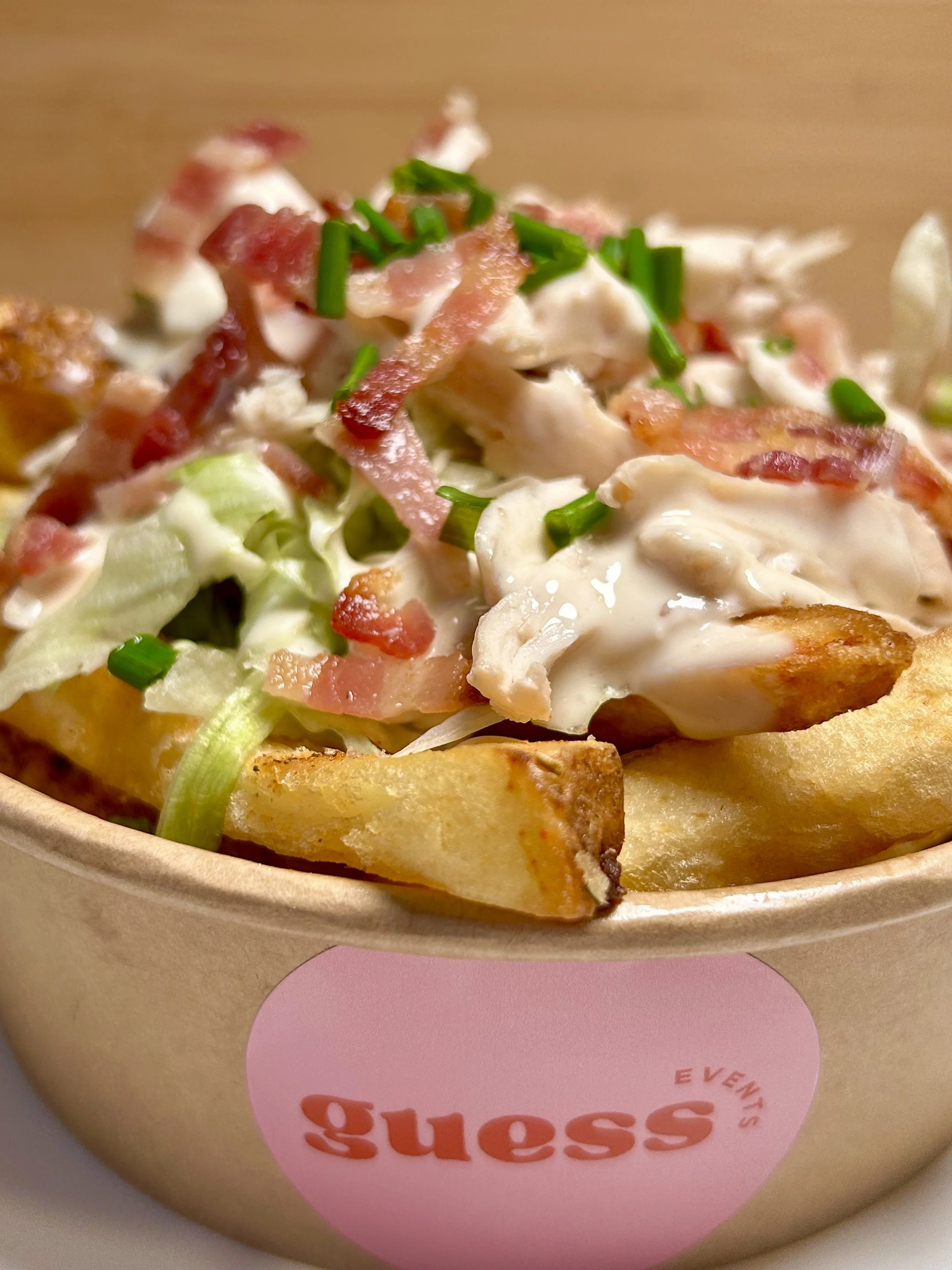A close-up image of a loaded fries with crispy bacon, chopped green onions, lettuce, and a caesar dressing in a paper container labelled with the guess events logo.