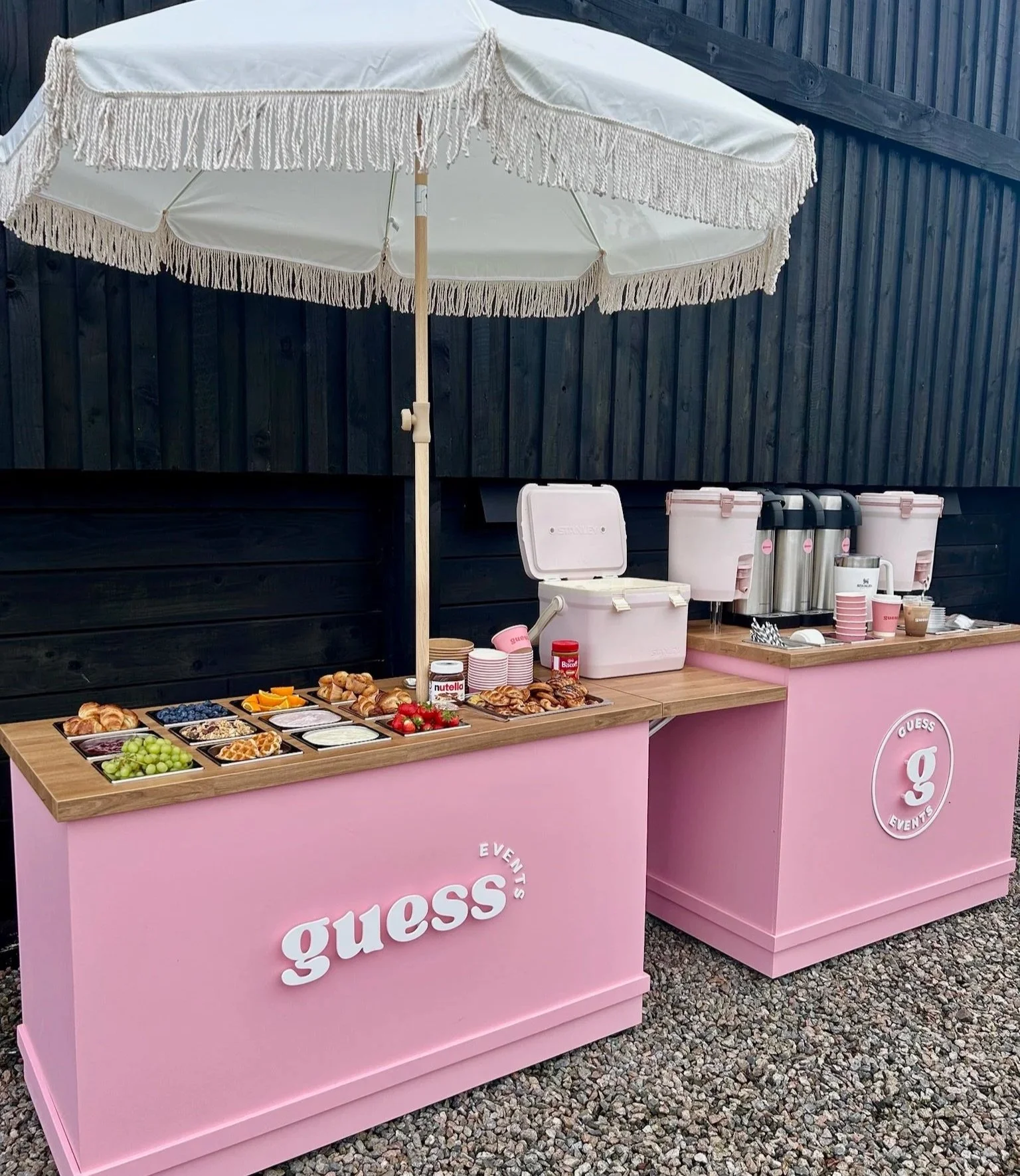 Outdoor snack station with pink counters, a white umbrella, and various breakfast items like croissants, grapes, and other snacks, with hot beverage dispensers in the background.