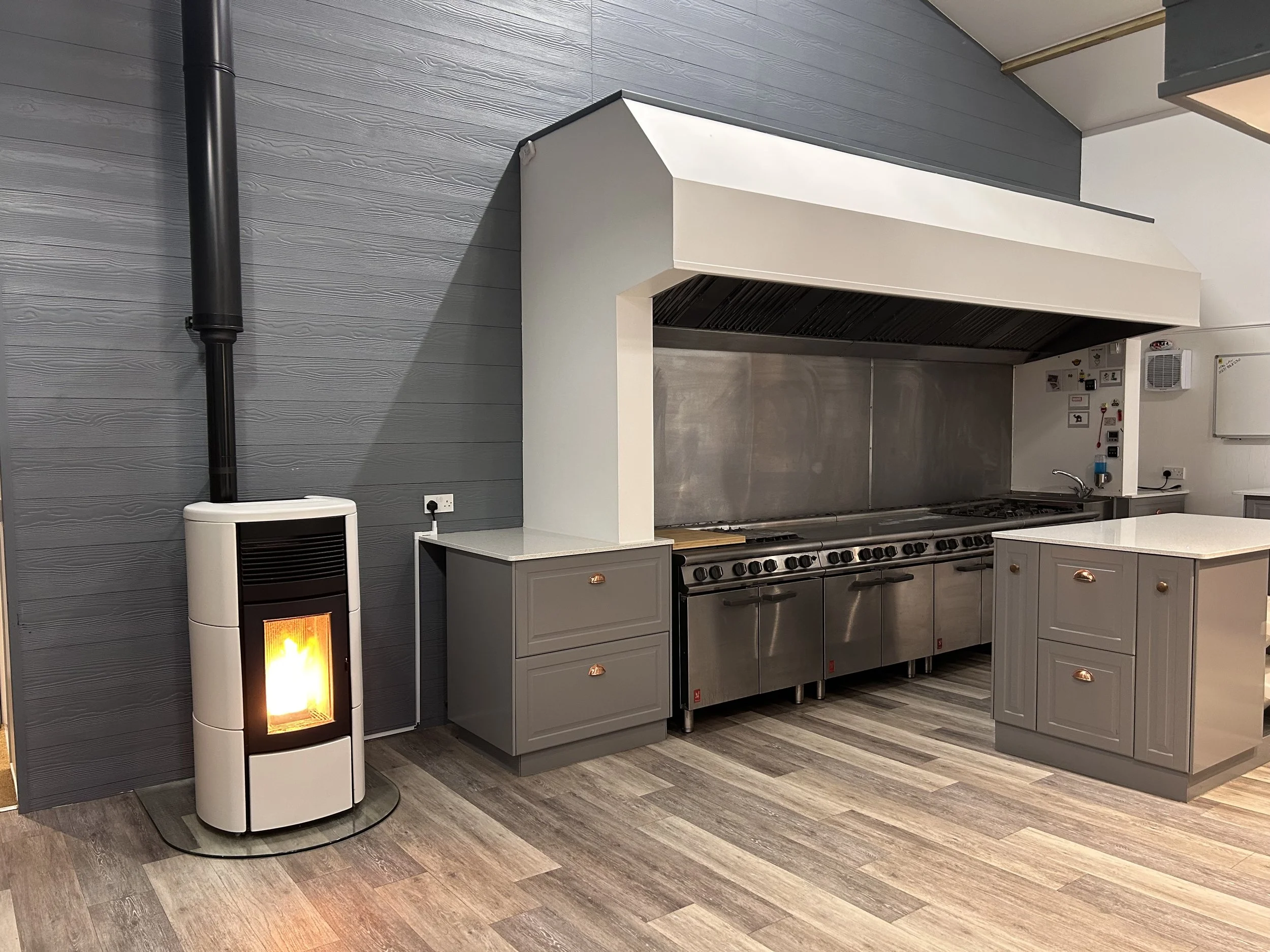 A commercial kitchen with gray cabinets, stainless steel appliances, and a wood-burning stove emitting orange flames. The kitchen has gray wooden walls and flooring, and a large stainless steel range hood above the stove.