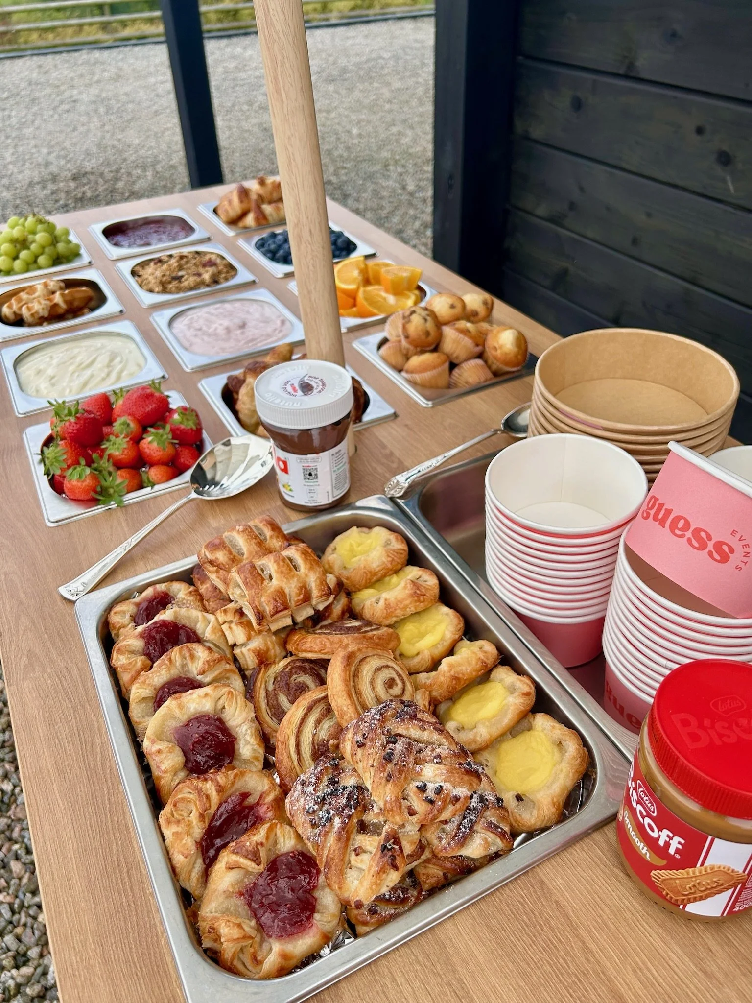 Assorted breakfast pastries, strawberries, blueberries, grapes, yogurt, whipped cream, fruit preserves, and small muffin cups on a buffet table outdoors.