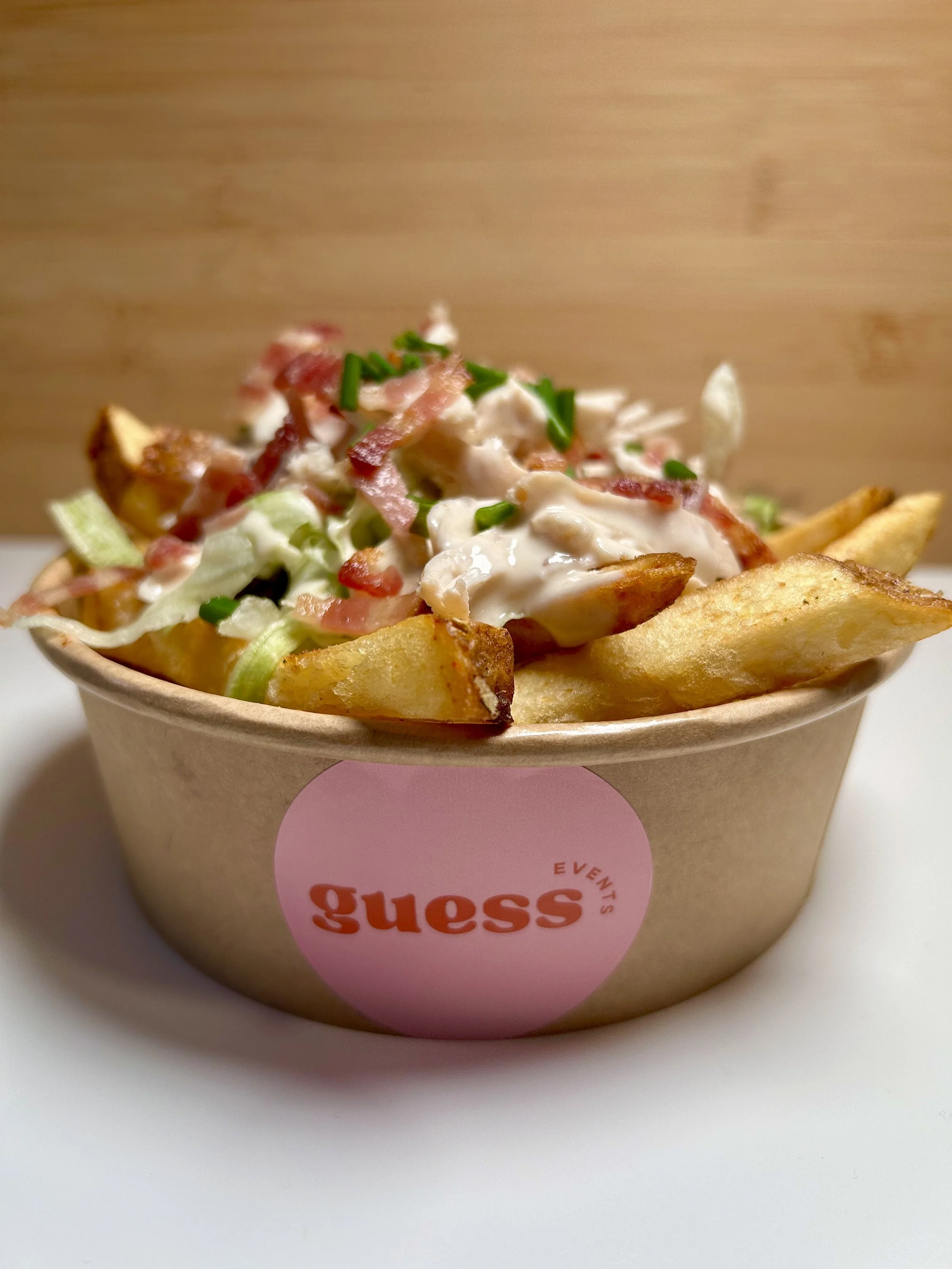 A bowl of seasoned French fries topped with bacon bits, shredded lettuce, chopped green onions, and creamy dressing, labeled with a pink and white "Guess Events" sticker.