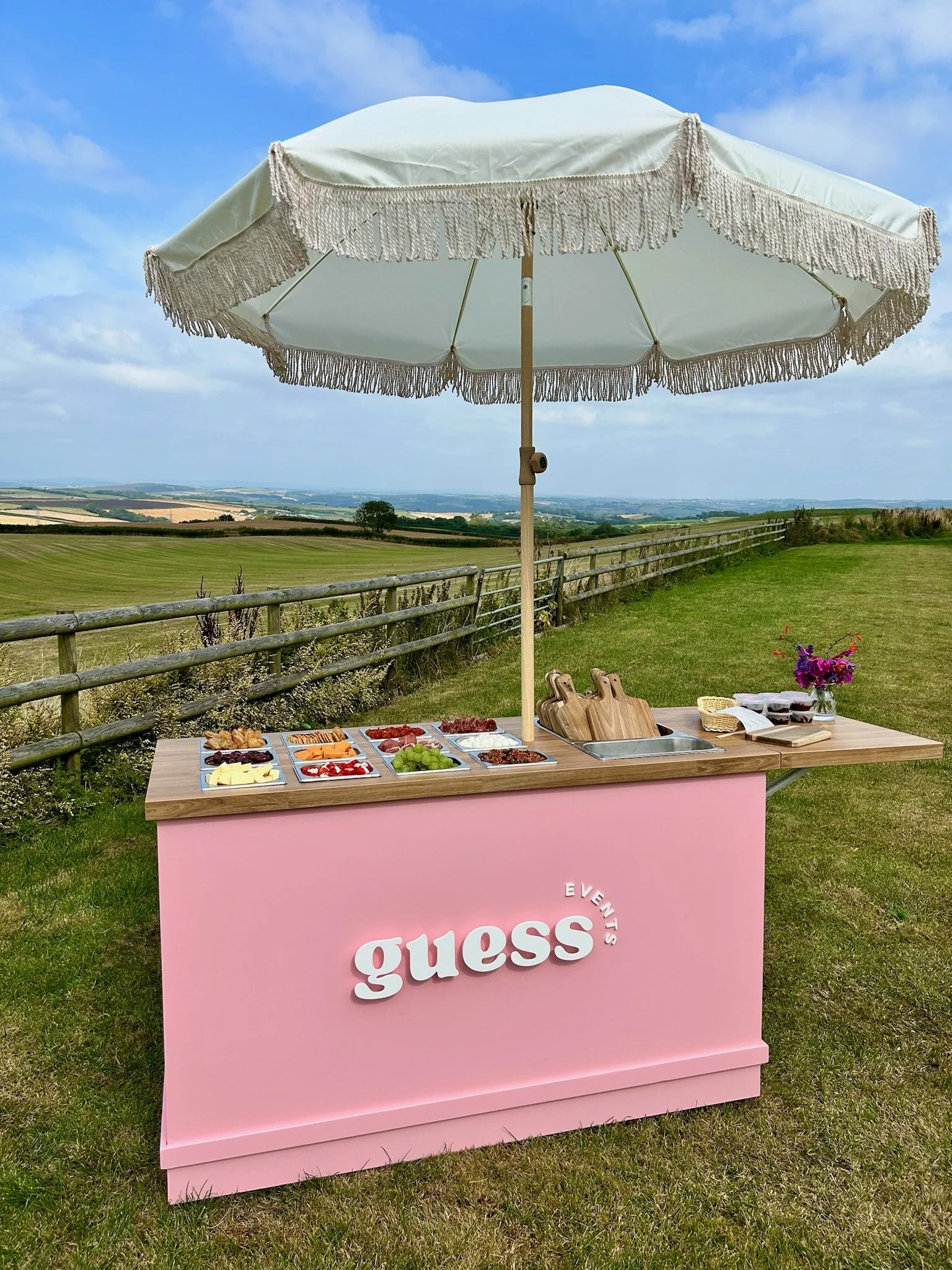 Outdoor buffet setup with a pink table labeled "guess," under a large white parasol, on a grassy area with a fence and open landscape in the background, featuring various food items, cutting boards, and a bouquet of flowers.