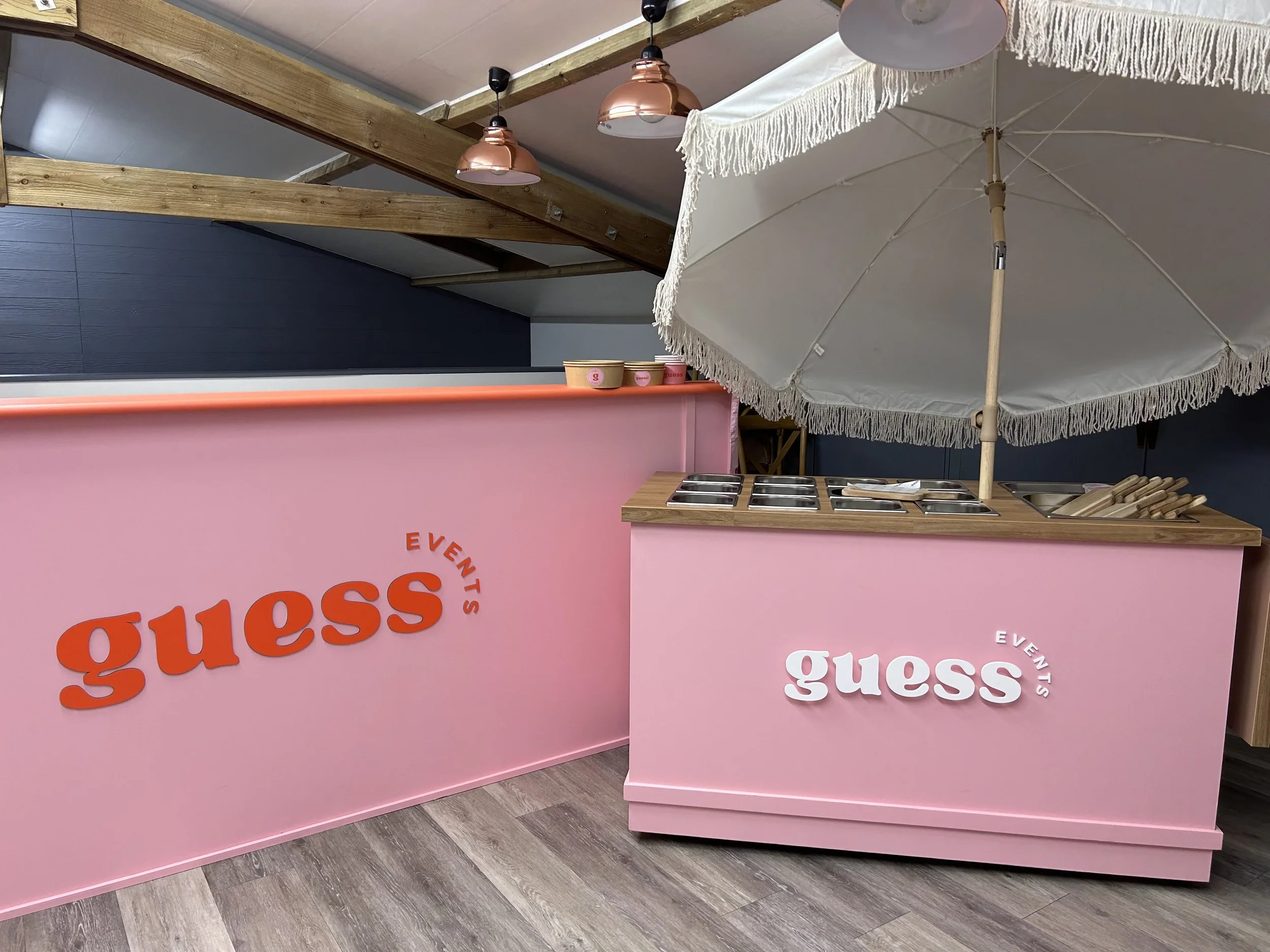 Pink snack bar with 'Guess Events' logo, ice cream or toppings station, white umbrella, wooden ceiling beams, and modern lighting fixtures.