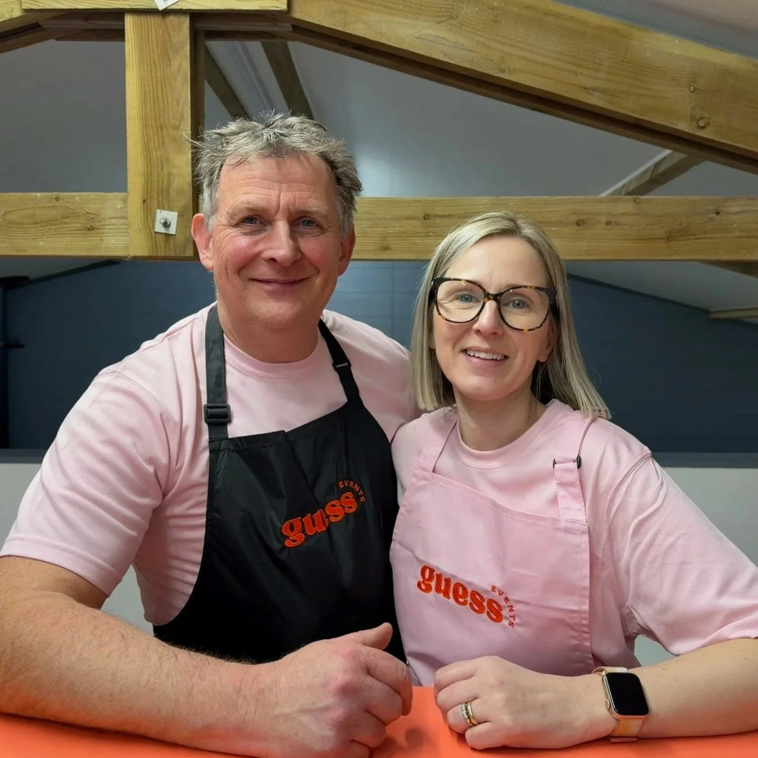 A man and woman, the owners of Guess Events wearing matching pink 'guess' shirts and aprons, smiling, standing close together, in a room with wooden beams in the background.