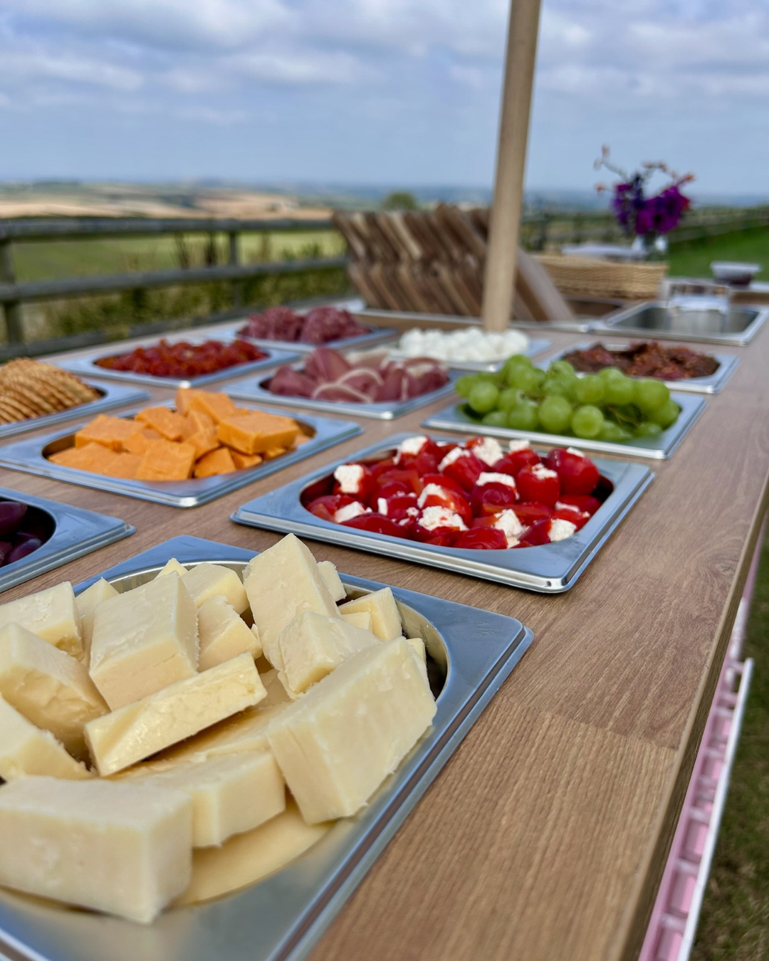 An outdoor spread of cheese, grapes, cherry tomatoes, cured meats, and other appetizers on a wooden table, with a scenic countryside view in the background.