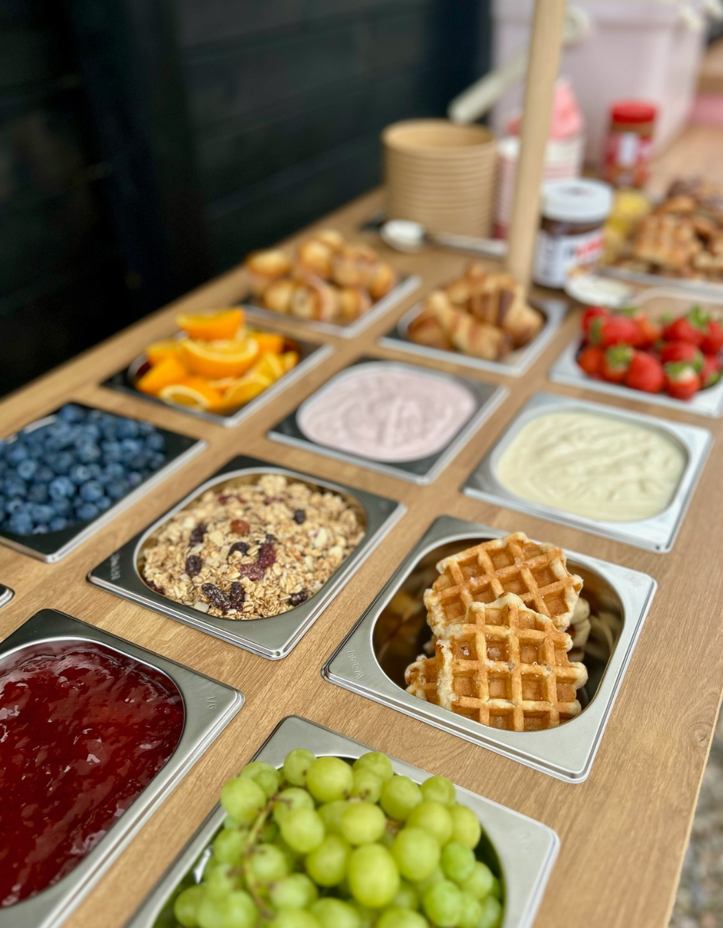 A buffet table with a variety of toppings including blueberries, grapes, sliced oranges, granola, strawberry yogurt, honey, strawberries, whipped cream, waffle halves, and various breakfast items.